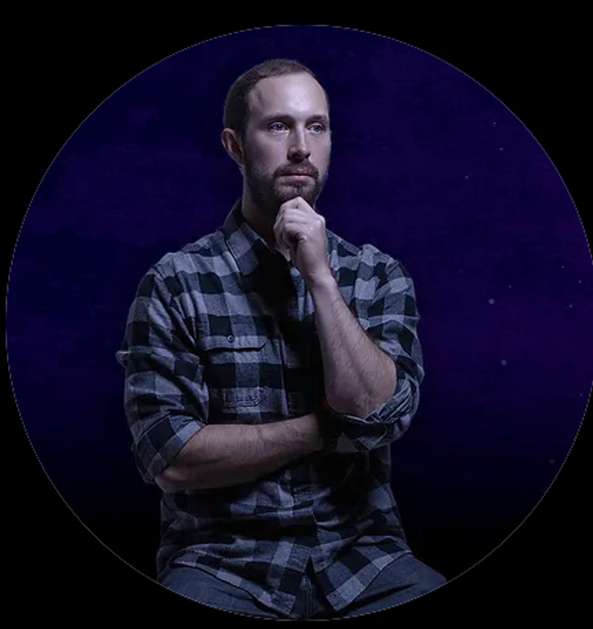 A white man in a flannel shirt. He is holding his hand to his chin, either in deep thought or to stop his head from falling off.