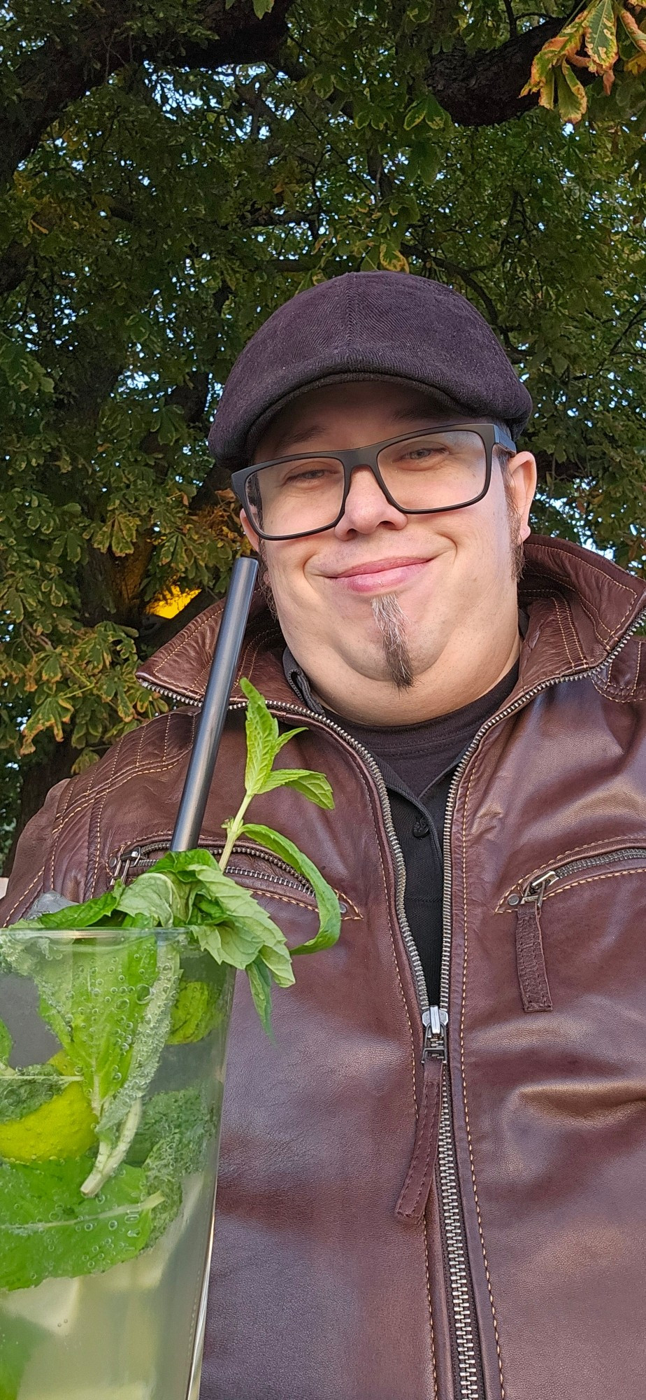 Typ mit schwarzer Brille, Koteletten und schmalem Kinnbart, trägt eine schwarze Schirmmütze und ein schwarzes Polohemd unter einer braunen Lederjacke. Im Vordergrund steht ein Mojito, im Hintergrund sind Bäume.