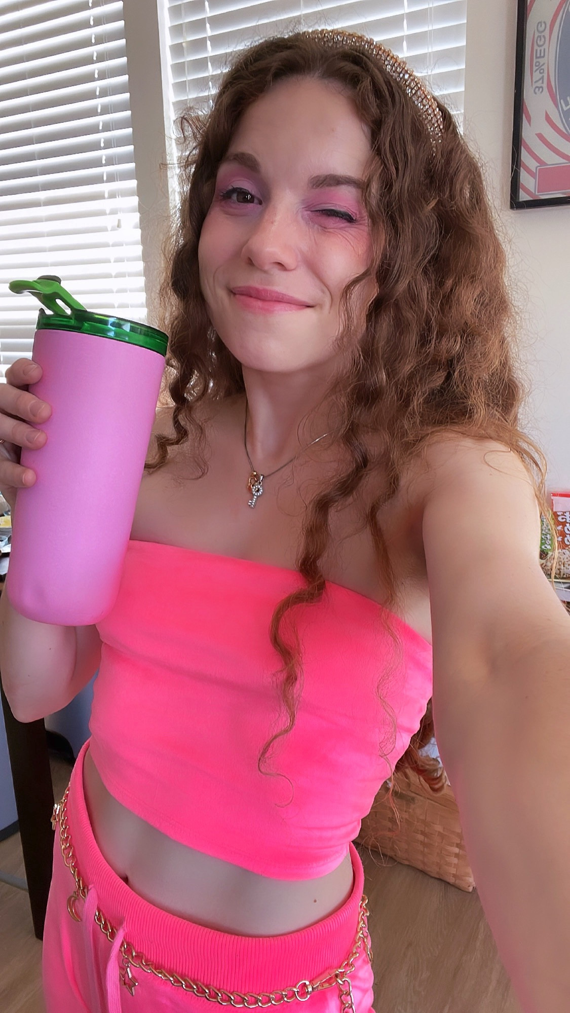 A shot of me winking at the front facing camera, wearing a bright pink tube top and and pants and holding a pink  and green tumbler of water.