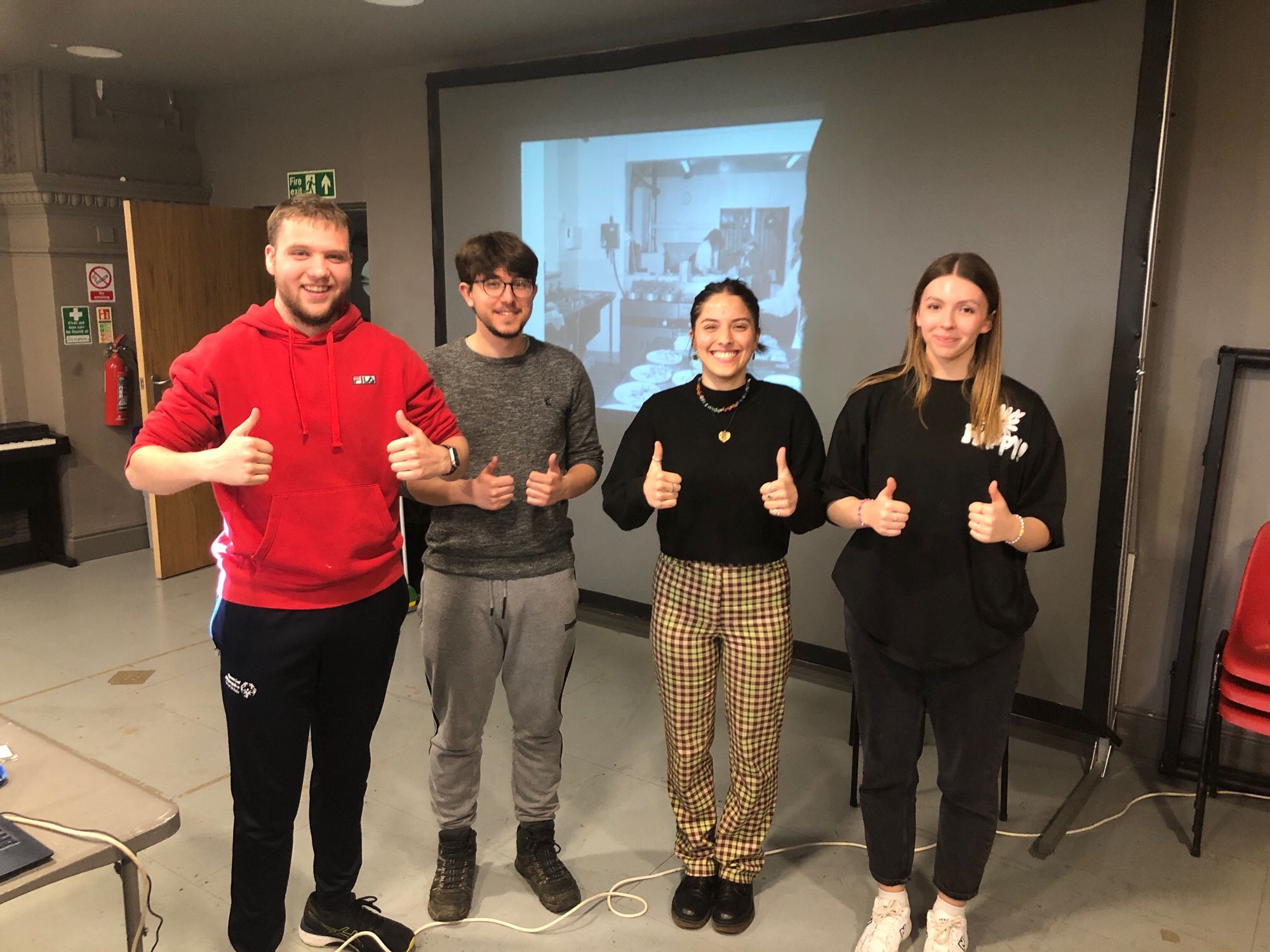 Four University of Huddersfield  students at  Pennine  Heritage. They are stood in front of a screen showing a black and white photo. They have their thumbs up and have very big smiles.