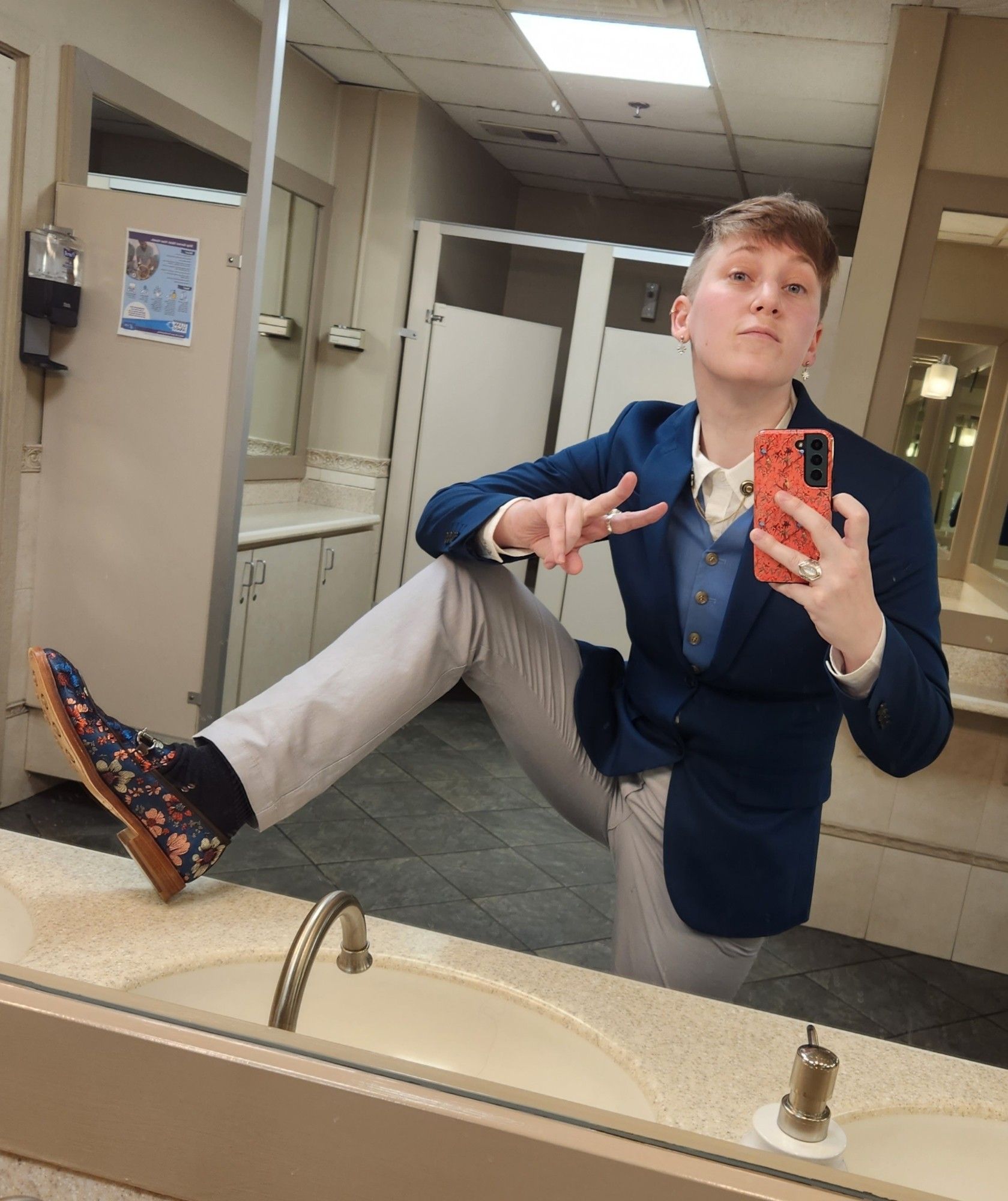 a person in gray trousers, a dark teal suit, a blue waistcoat, and teal floral shoes posing in front of a bathroom mirror of a wedding venue. their heel is somewhat rudely propped onto the bathroom sink in order to get the whole outfit in, however the surface was wiped down afterward. they are giving a peace sign to the camera because they are bisexual and that's kind of a go-to.