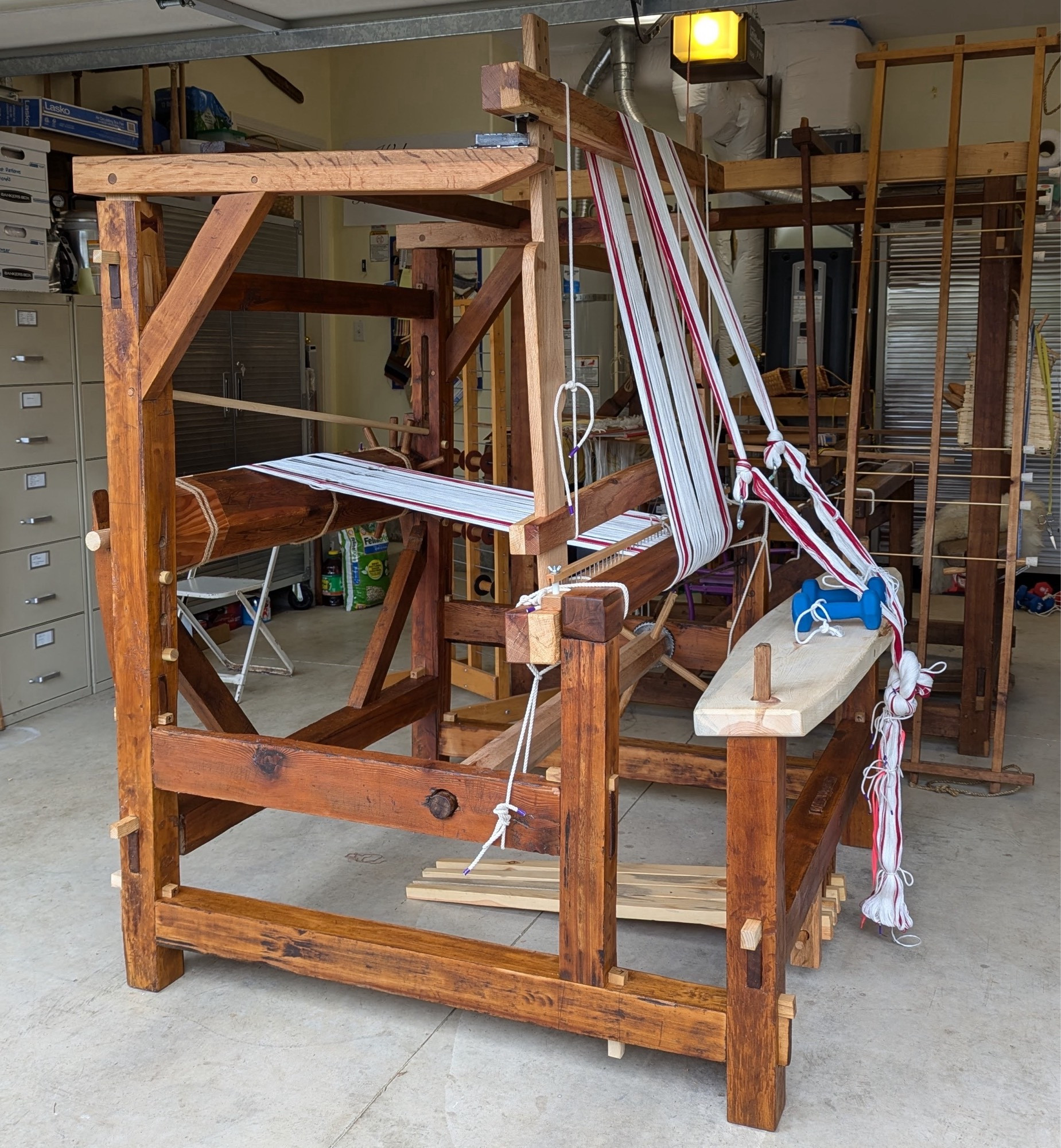 A side view of an antique cantilevered hand loom, in the process of have a short warp of white and red towels beamed on.