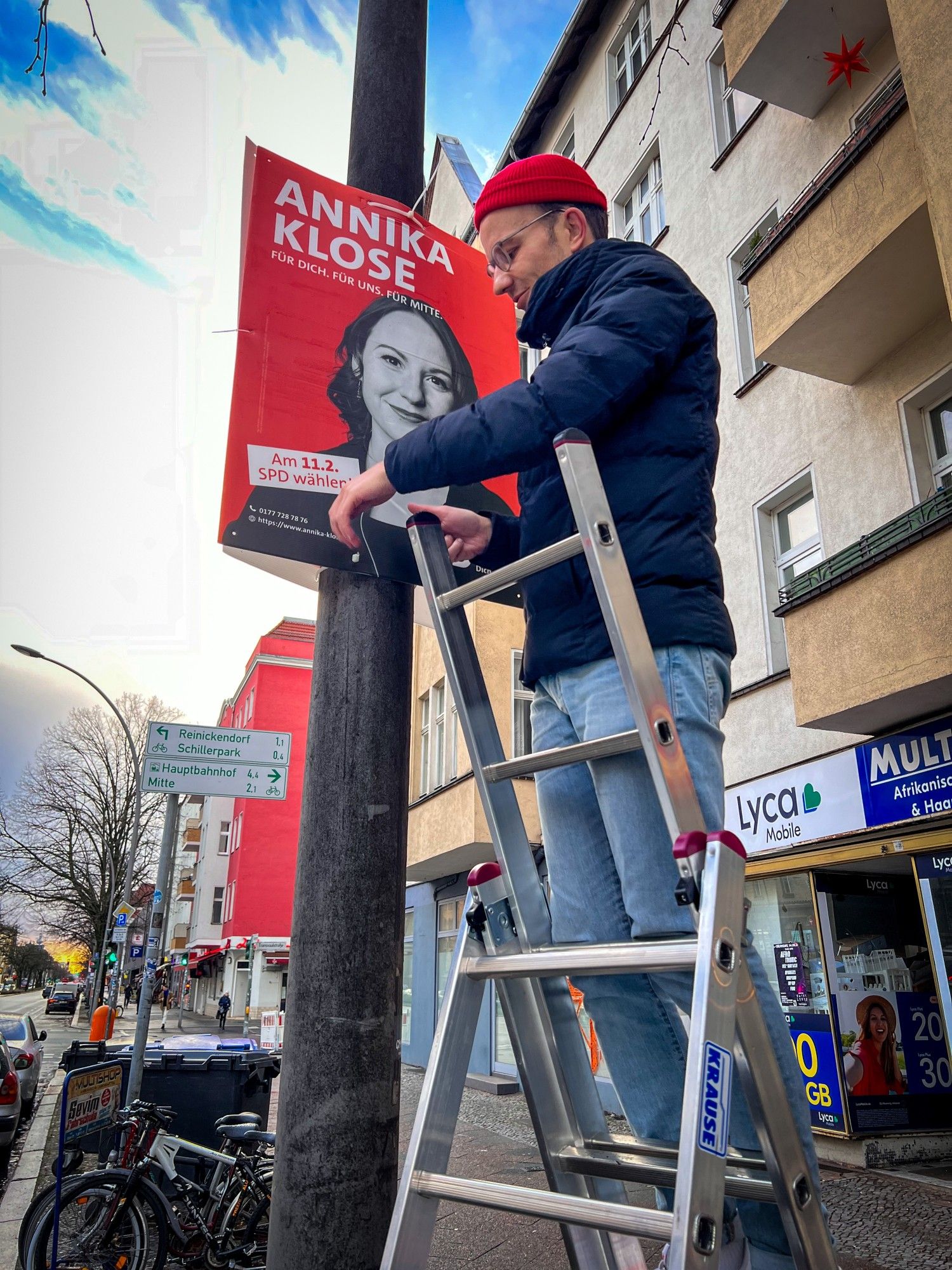 Auf der Leiter stehend beim Plakate aufhängen. Zu sehen ist ein Plakat der Bundestagsabgeordneten der SPD für Berlin Mitte, Annika Klose. Im Hintergrund sind die Wohnhäuser entlang der Müllerstraße zu sehen.