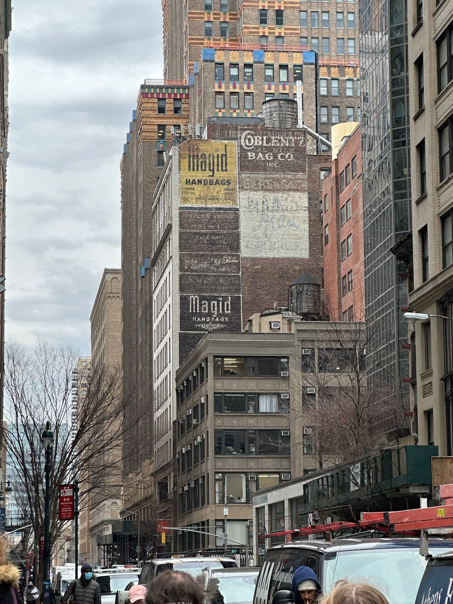 Photo up a street of New York showing multicolored buildings, a couple of probably no longer used water tanks on roofs, and some faded advertisements painted on the blank brick side of a tall old building, advertising Magid Handbags and the Coblentz Handbag Co.