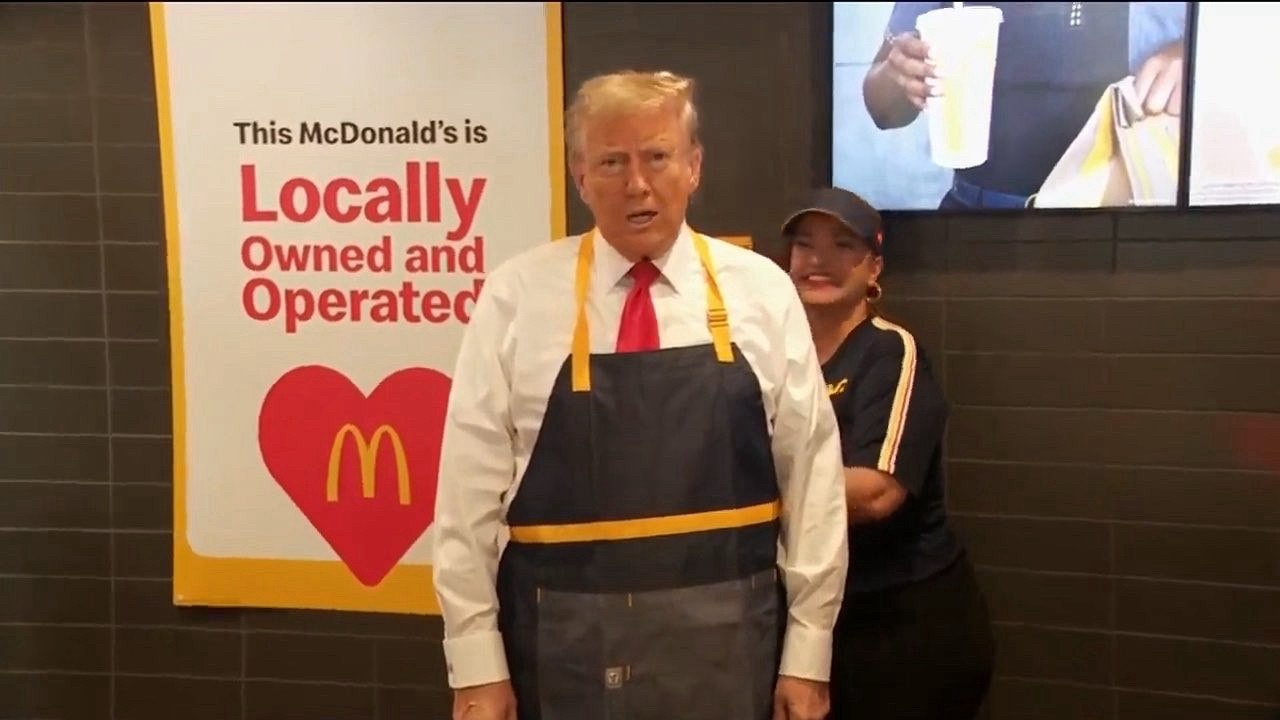 A grinning McDonad's employee ties an apron around Donald Trump's midsection. Trump is staring forward with cold, lifeless eyes. His gaze comes from deep within the prison of his own failing flesh. There is a sense of dull horror in his posture, perhaps the handful of functional neurons still firing realize that nothing can save him from soon returning to the dust we are all molded from. There is no escape from this, regardless of the election. The shadows grow deeper and closer every day and have pushed the light from his eyes.

Also his hair is fucked up and stupid.