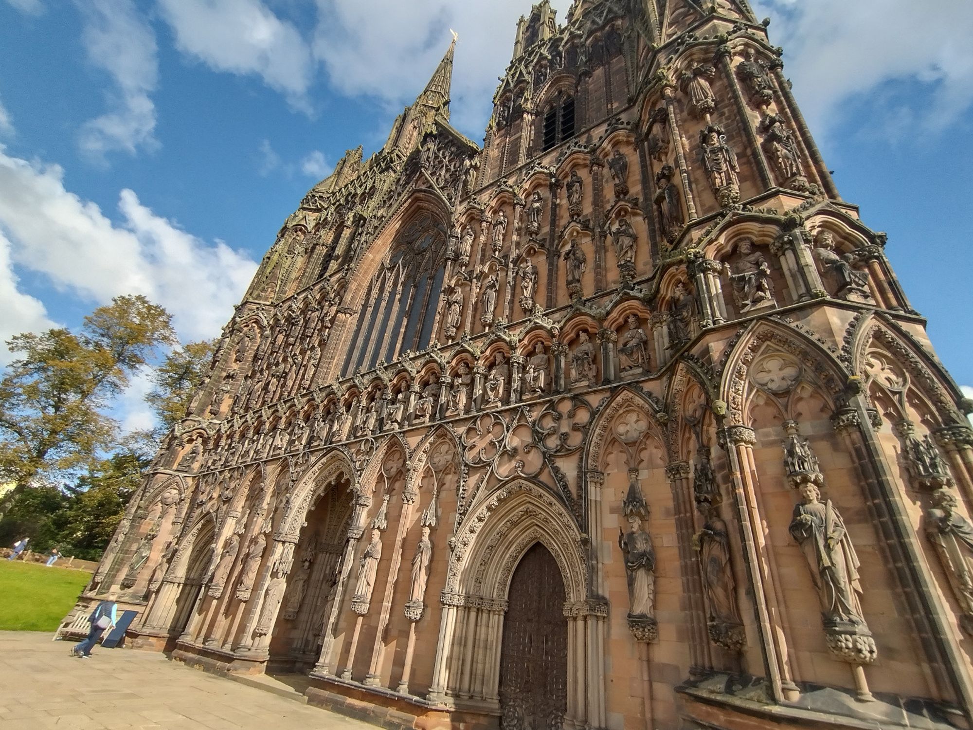 A photograph of the west face of Lichfield Cathedral, resplendent in brown stone blocks, gothic arches, and numerous statues of different sizes depicting kings, queens, and saints. The sun is just catching the side of the carvings.