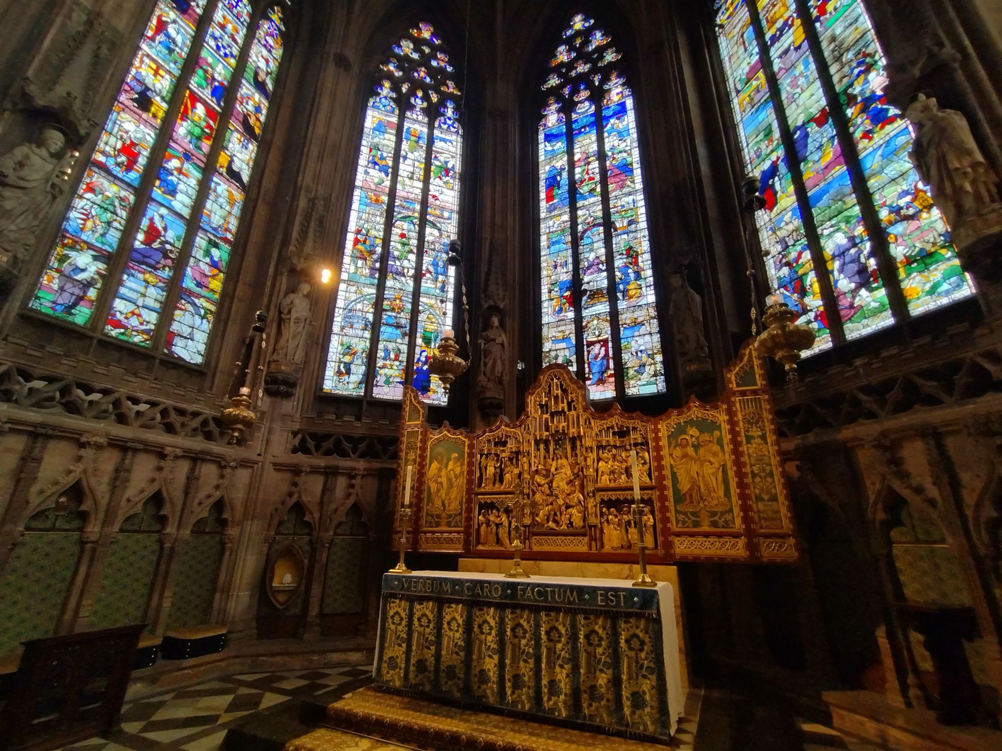 A photograph of Lichfield Cathedral high altar. The chancery is surrounded by high gothic arched windows filled with colourful art depicting biblical scenes. Behind the altar is a gold freeze, filled with more details. The front of the altar is decorated with a vertical pattern. The plainest thing in the scene is the white cloth over the altar with two tall shiny candlestick holders resting on it.