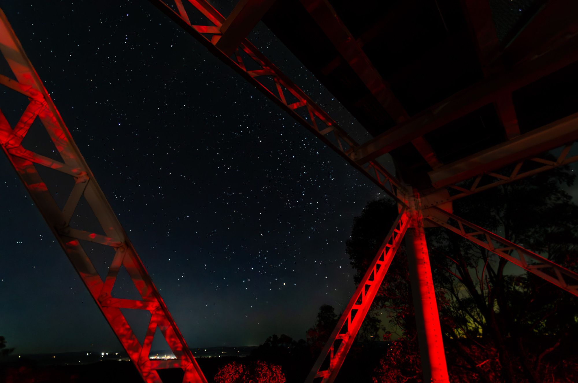 The night sky through the girders of a metal fire watchtower. The sky is almost fully dark with brilliant stars; the metal girders are illuminated red by brake lights from the parking lot a floor down and 200 metres away.