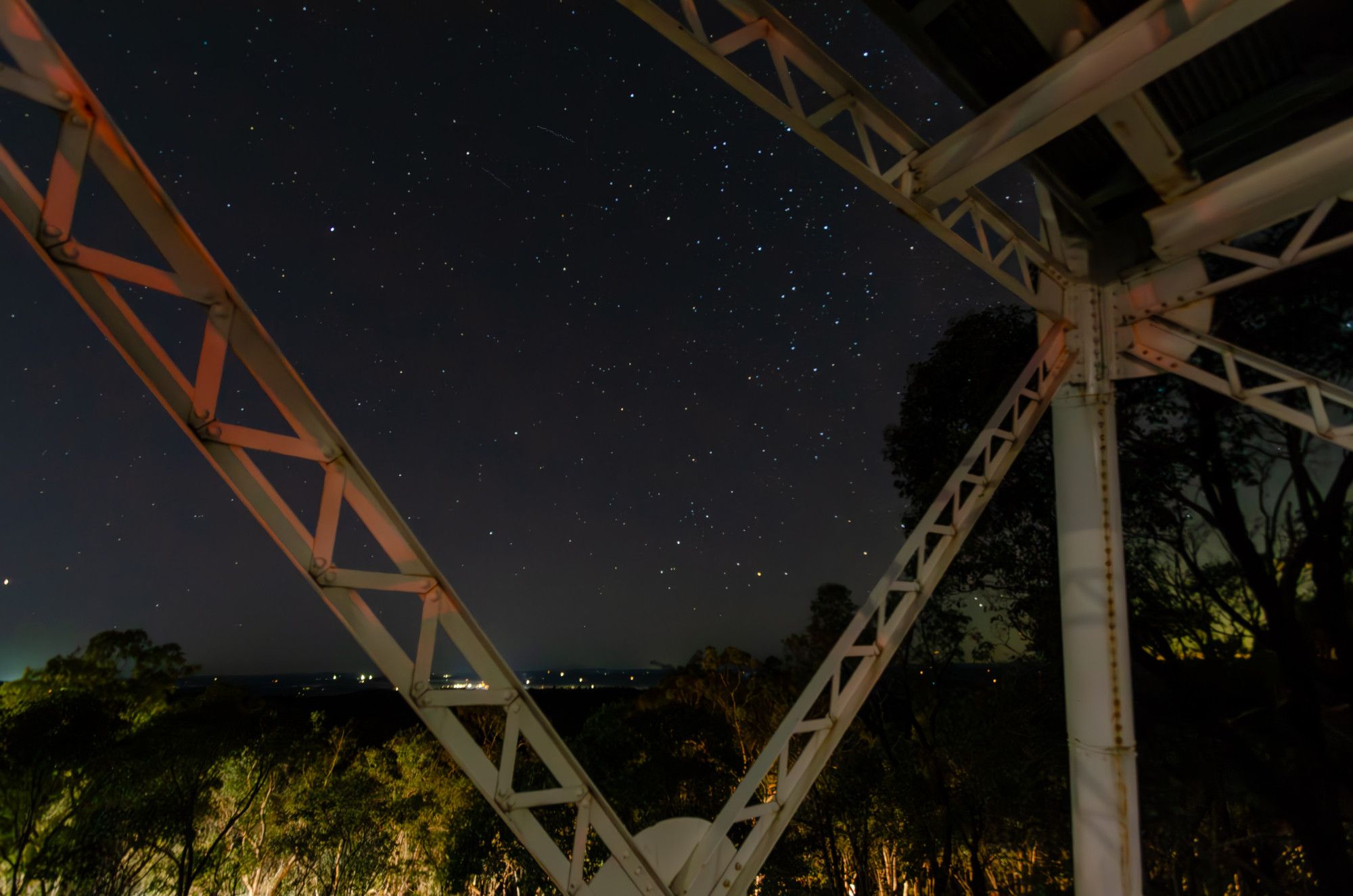 A view of the night sky through the metal girders of a fire watchtower. The trees in the foreground are lit by the headlights of a car driving through them, and there's still a little light in the sky, but the stars are clearly visible.