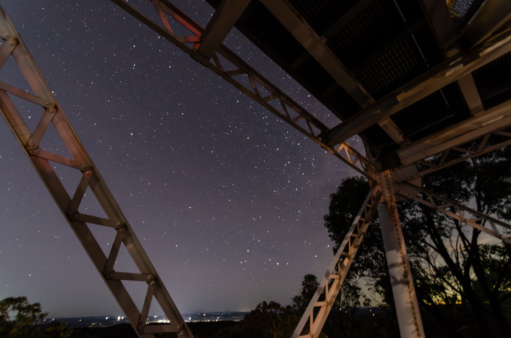 View of the night sky through a metal fire watch tower structure. It's past dusk but the sky is reflecting some light from towns and the last of sunset.