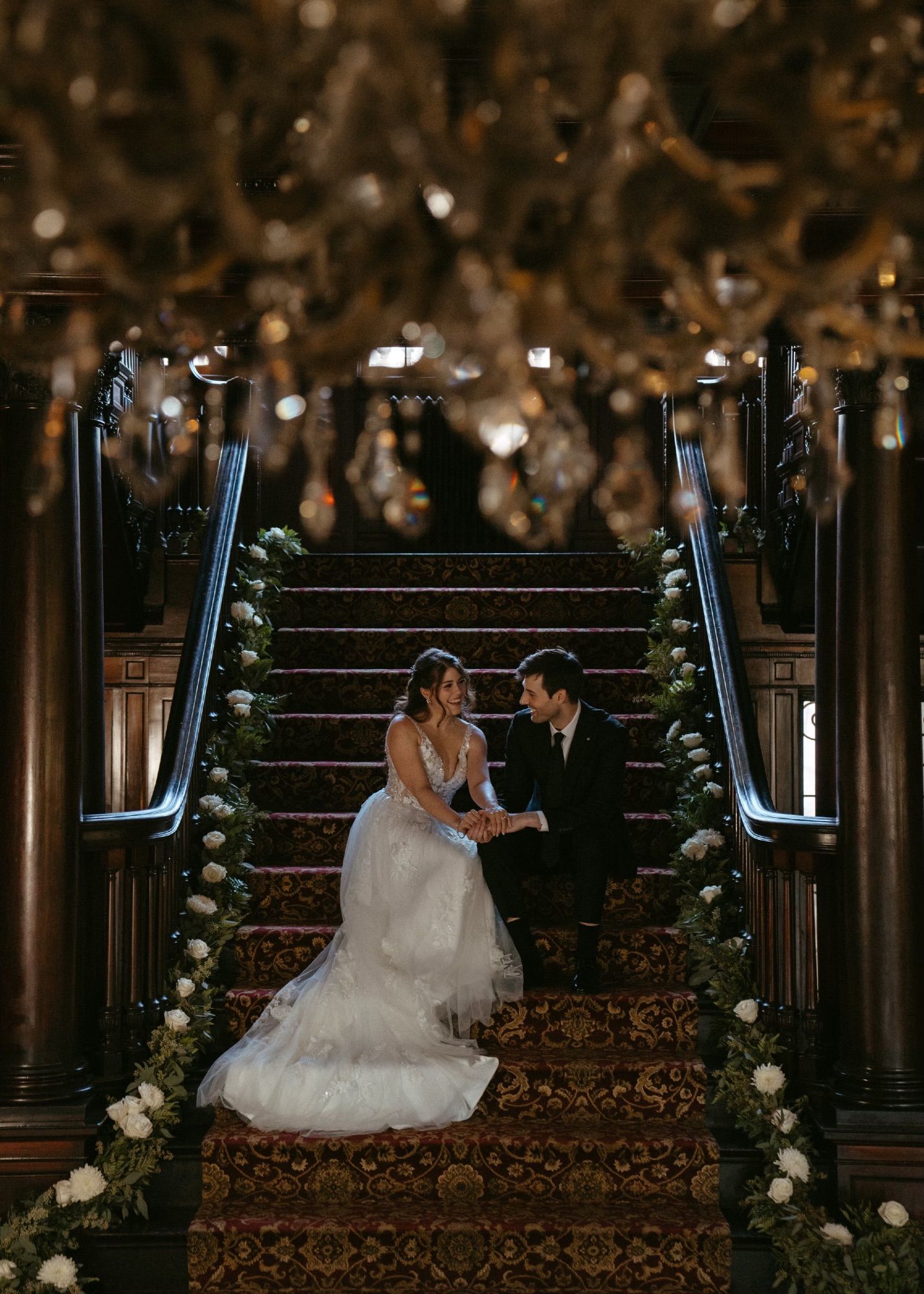 Staircase in opulent mansion lined with eucalyptus. Me and my partner Anna in formal wear sitting down on the steps smiling. A chandelier frames the  top of the shot with dangling crystals. 