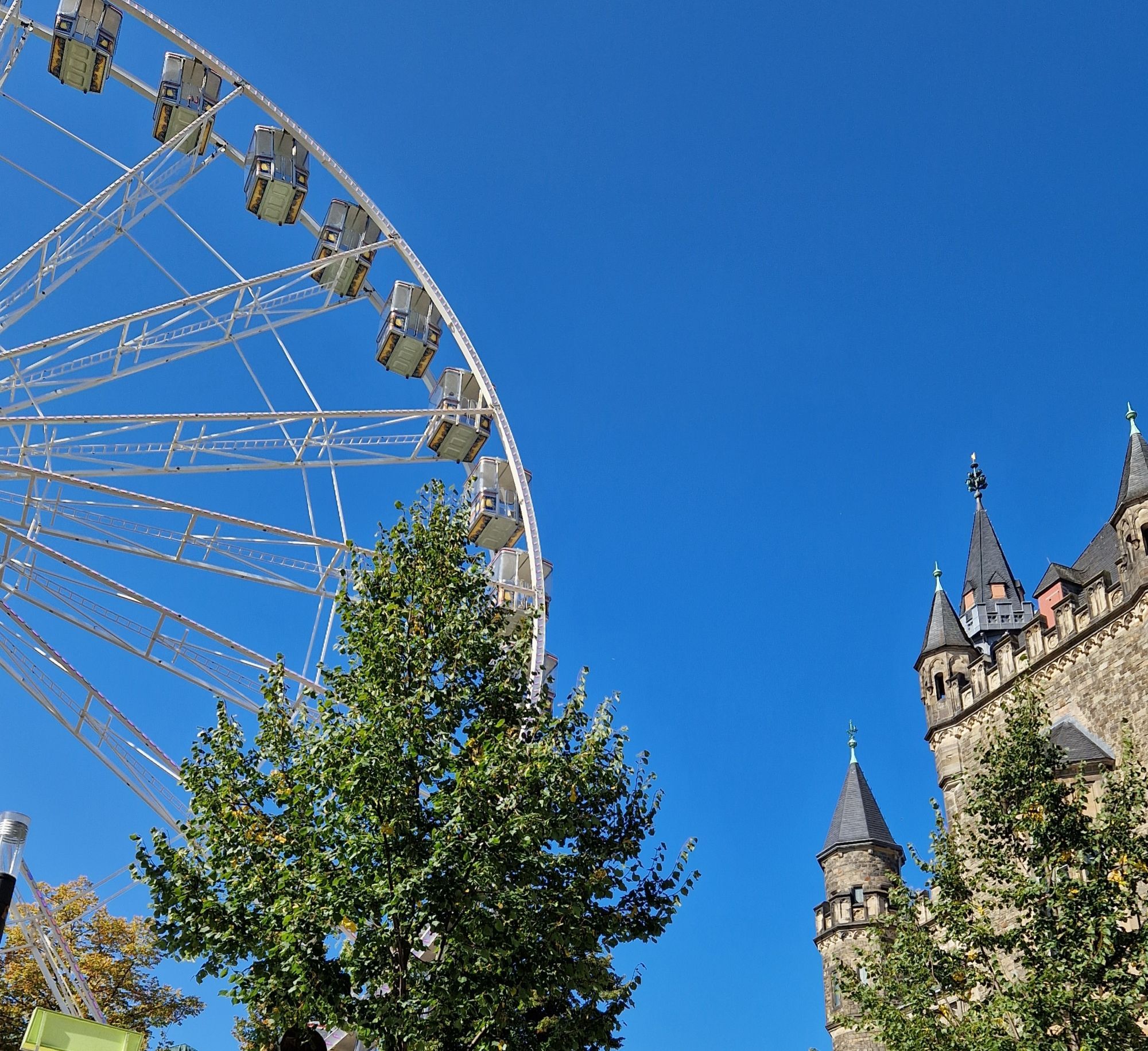 Vor blauem Himmel steht links ein Riesenrad. Umrahmt von Bäumen Auf der rechten Seite ist ein Teil des Aachener Rathauses zu sehen.