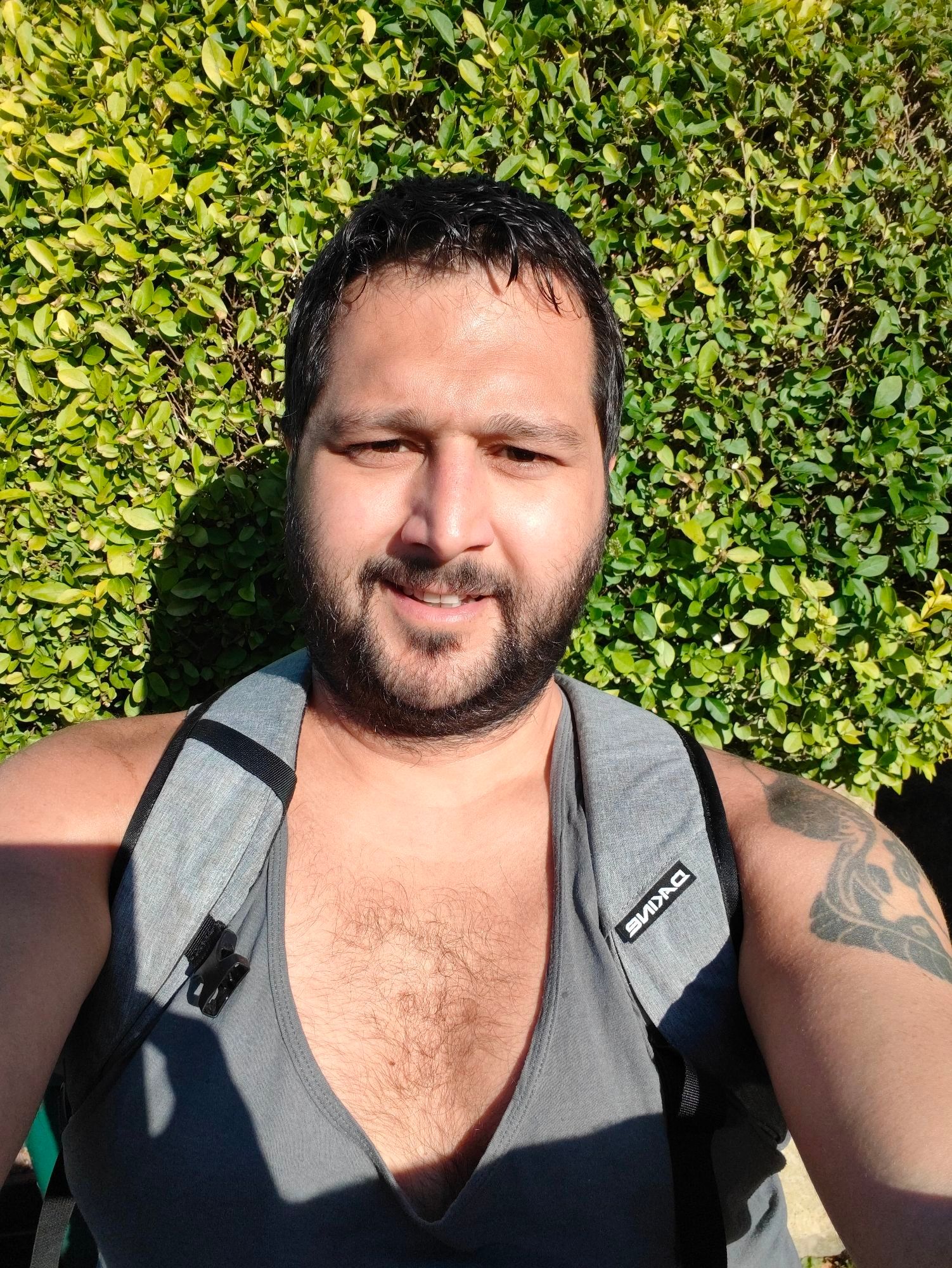 Me with short but messy black hair staring into the camera wearing a grey rucksack and charcoal running tank vest with my arms, shoulders and chest exposed. I am standing in front of a leafy green bush