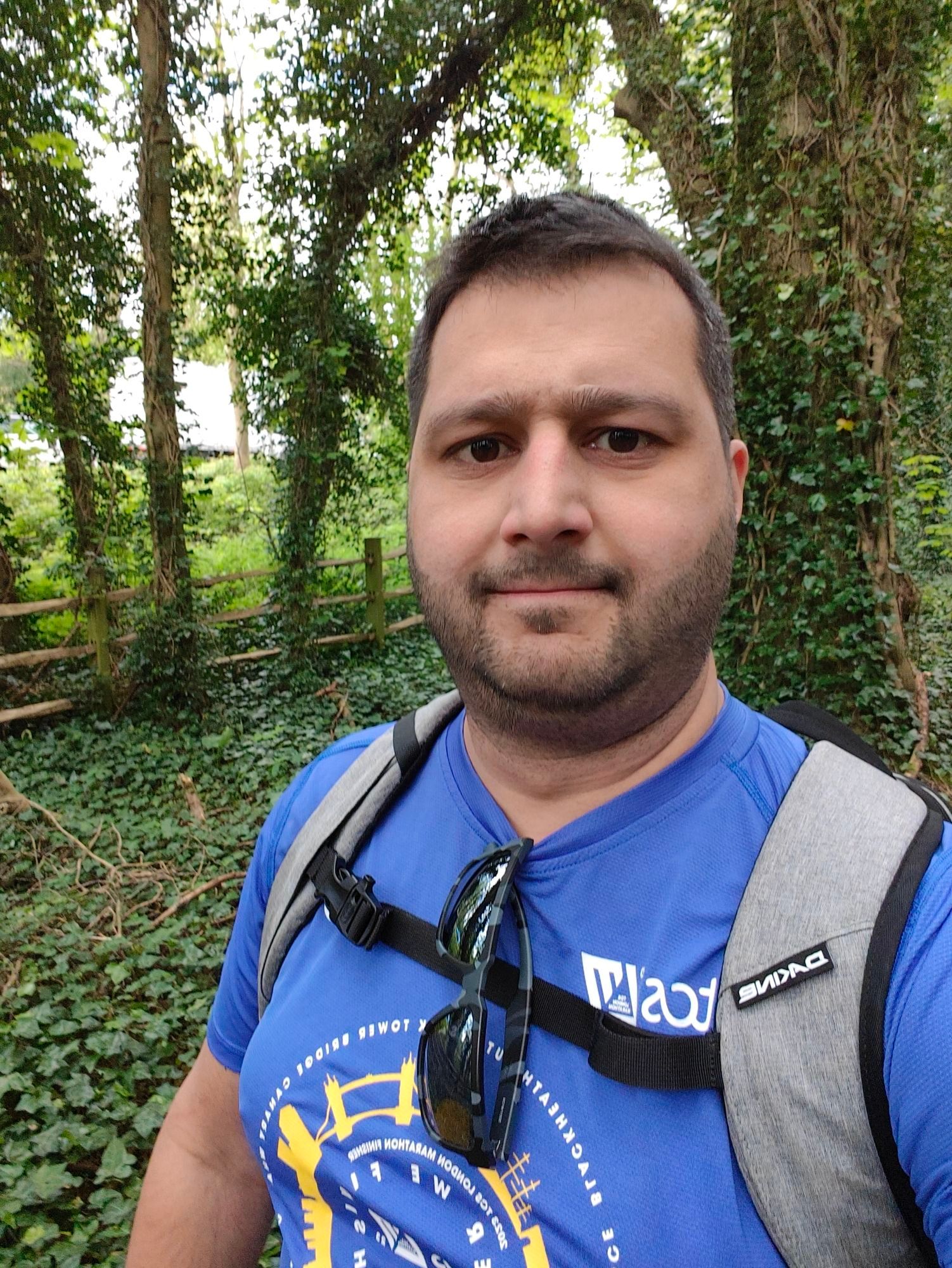 Me wearing my blue 2023 London Marathon finishers running top and a grey rucksack, staring into the camera when in a green wooded area