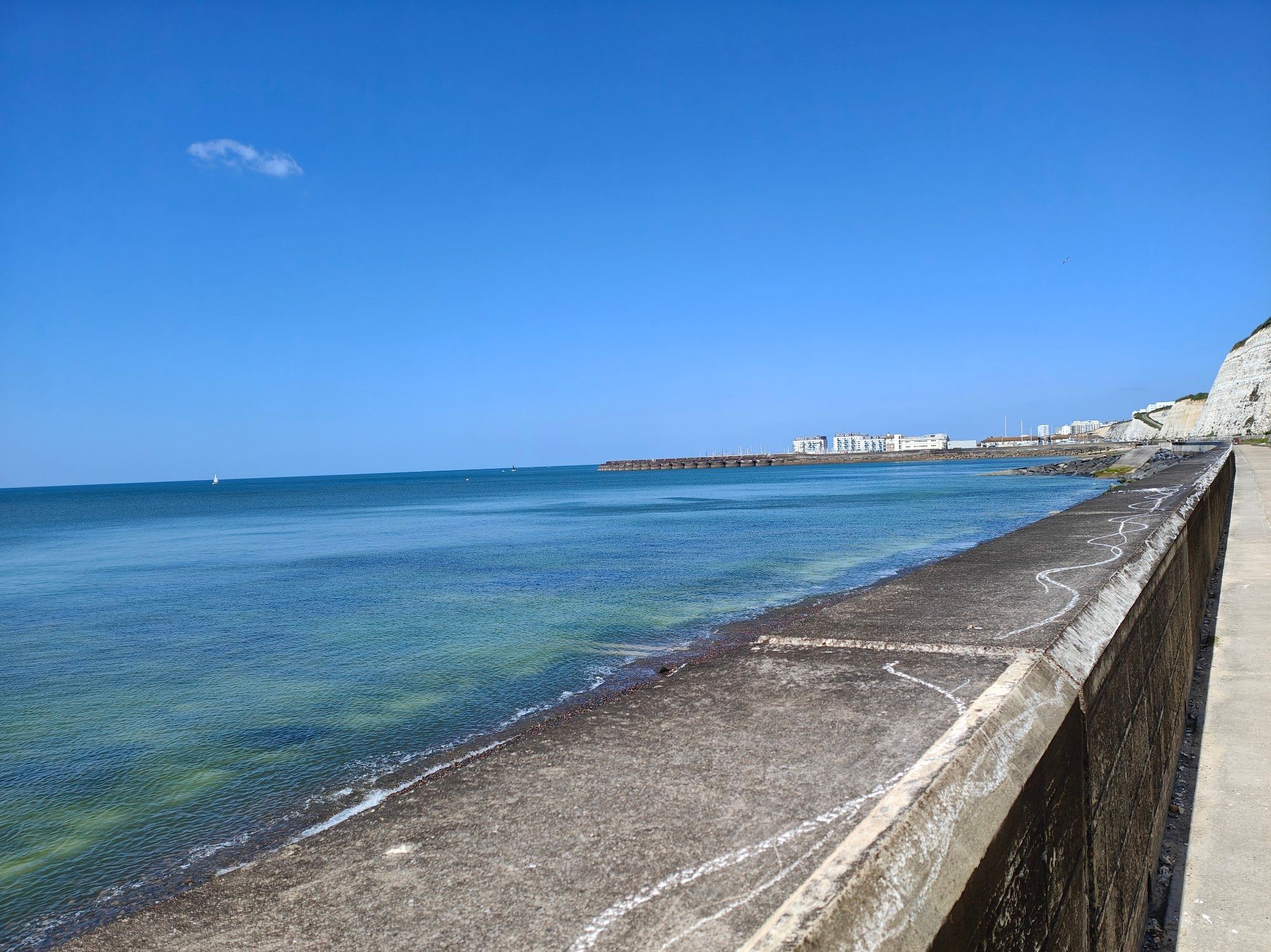 Blue sea with white chalk from the rock pool underneath. A concrete wall and path is visible, from where the photo was taken. The sky is blue and no cloud