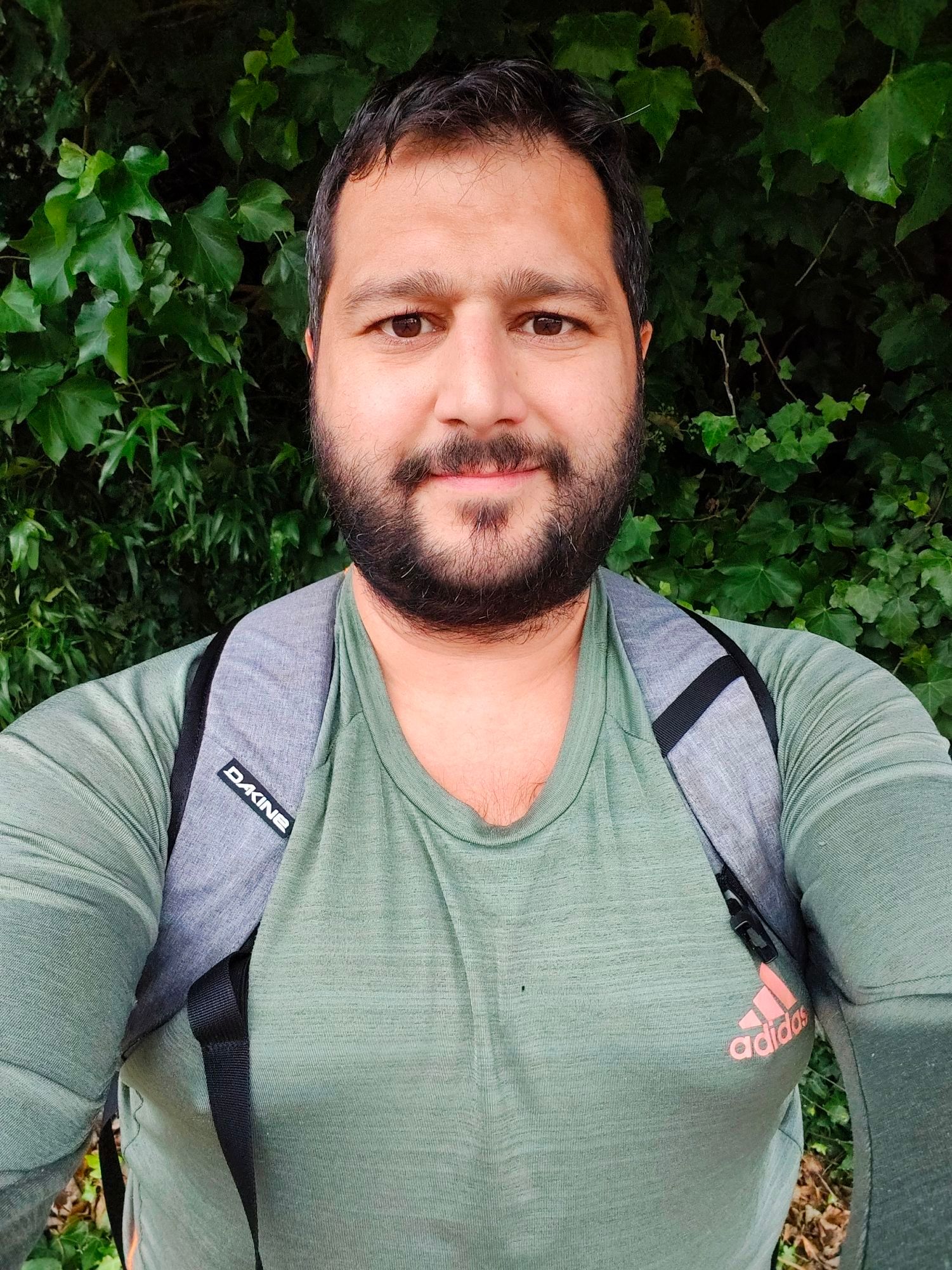 An olive skinned man with a beard (me) is staring at the camera in a long sleeve olive green running top. The man stands in front of a green bush wearing a grey rucksack.