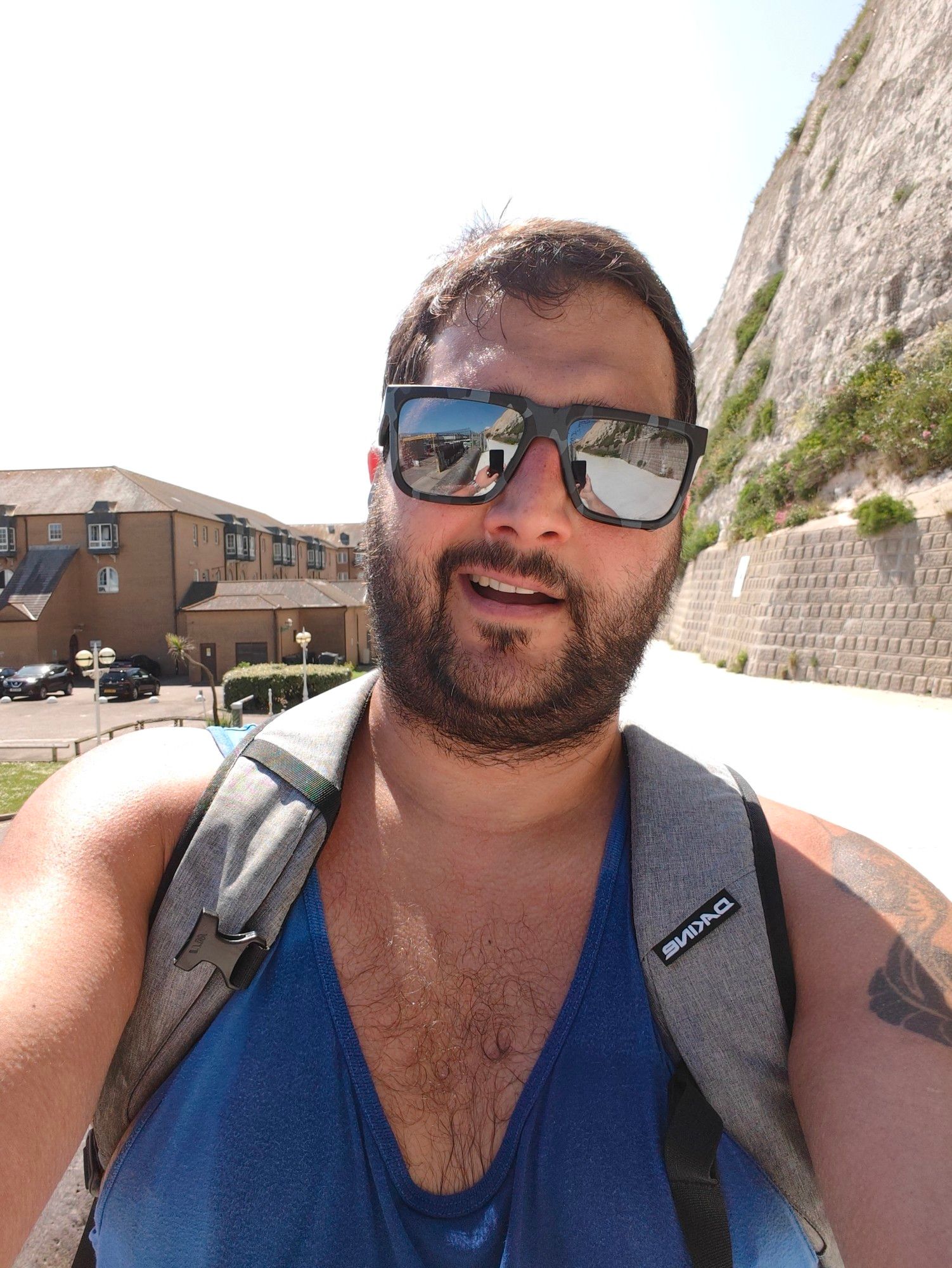 A bearded,  brown skinned man with a beard wearing sunglasses, a blue running top and grey rucksack looks tired but smiles to the camera on a concrete path. The man is me