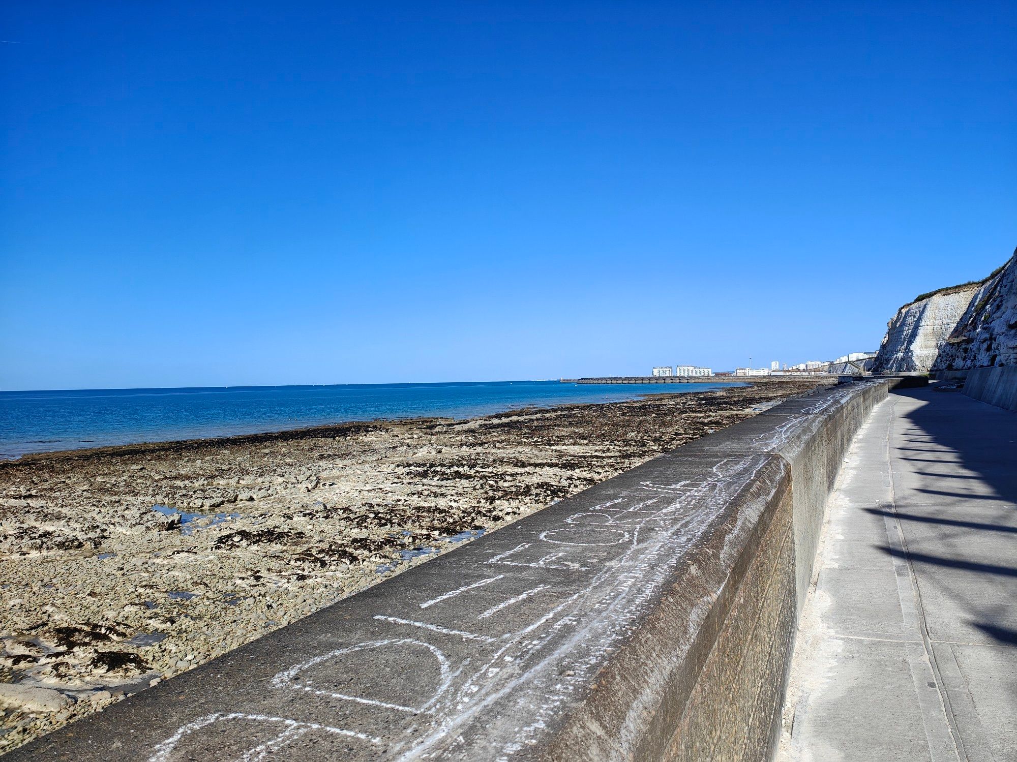 A concrete path. To the left a concrete wall that follows the path is visible with pebble and rock beach to the left of the wall. The blue sea can be seen to the far left adjoining the beach. To the right are white cliffs