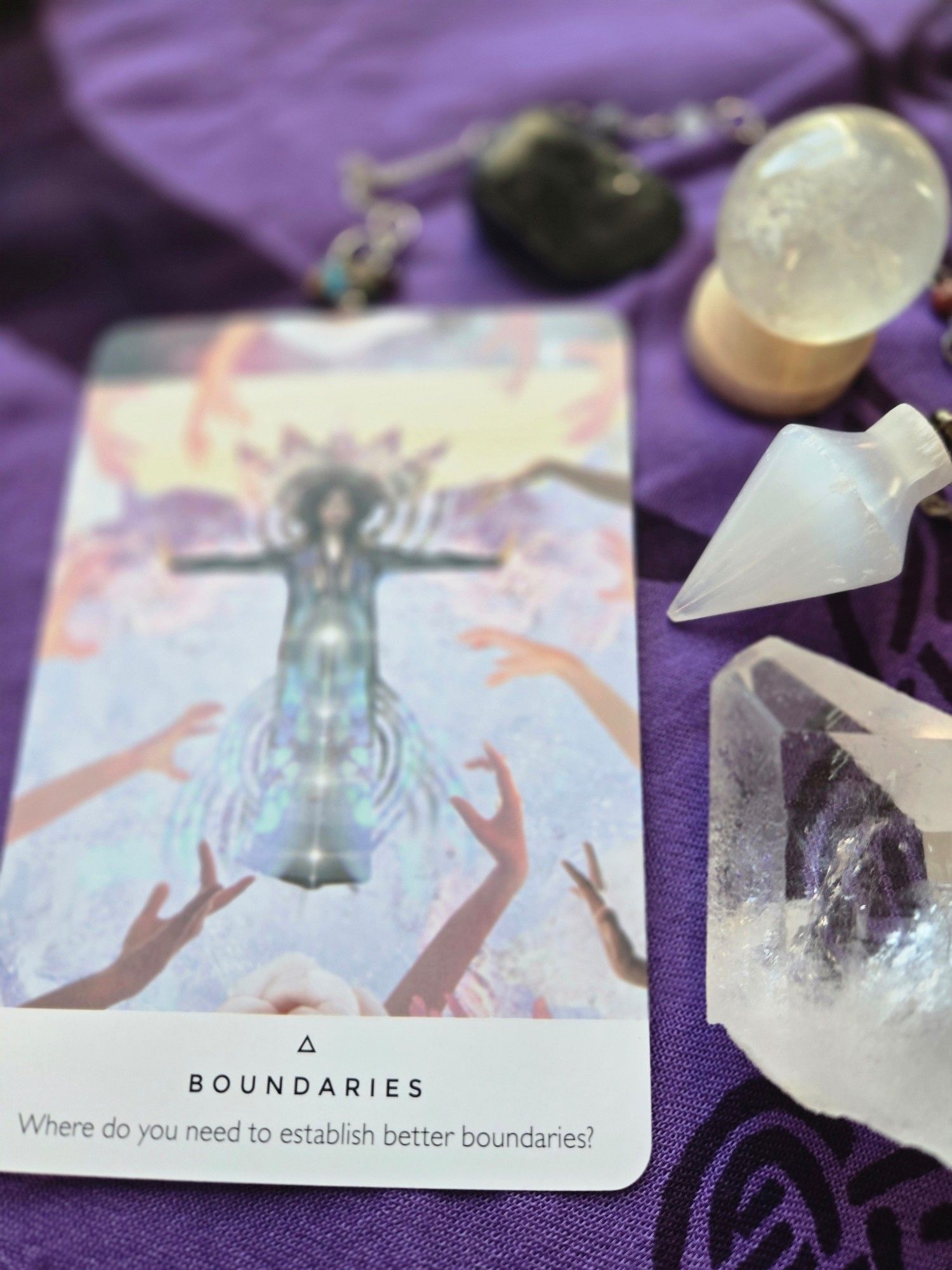 An oracle card on a purple and black cloth, surrounded by crystals and a pendulum.