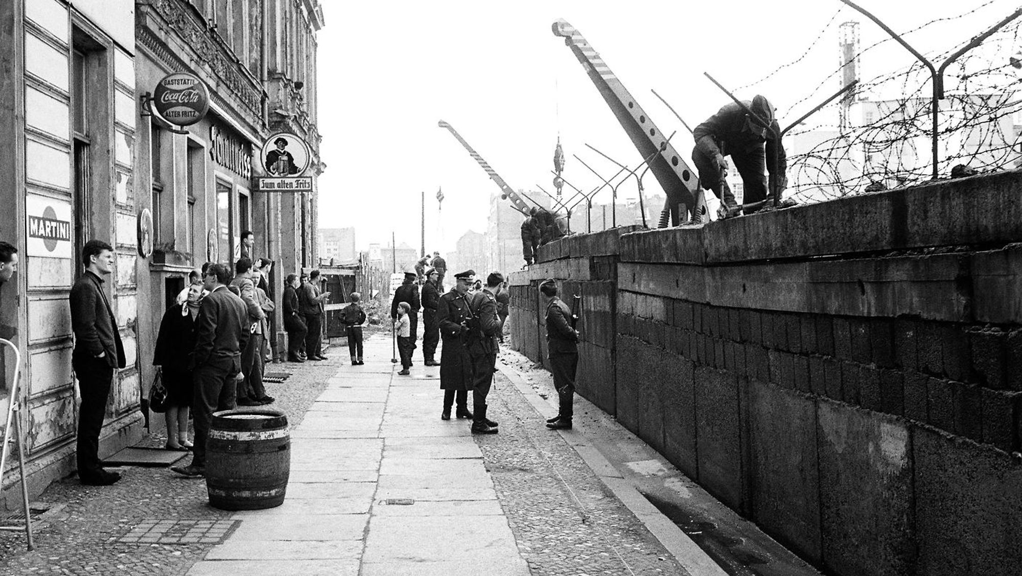 Foto vom Mauerbau - sie zerschneidet eine innerstädtische Straße, Westberliner schauen zu, wie die Mauer errichtet wird, vor der Mauer auf der Westseite stehen DDR-Grenzer, weil ein Meter noch zu Ost-Berlin gehörte.