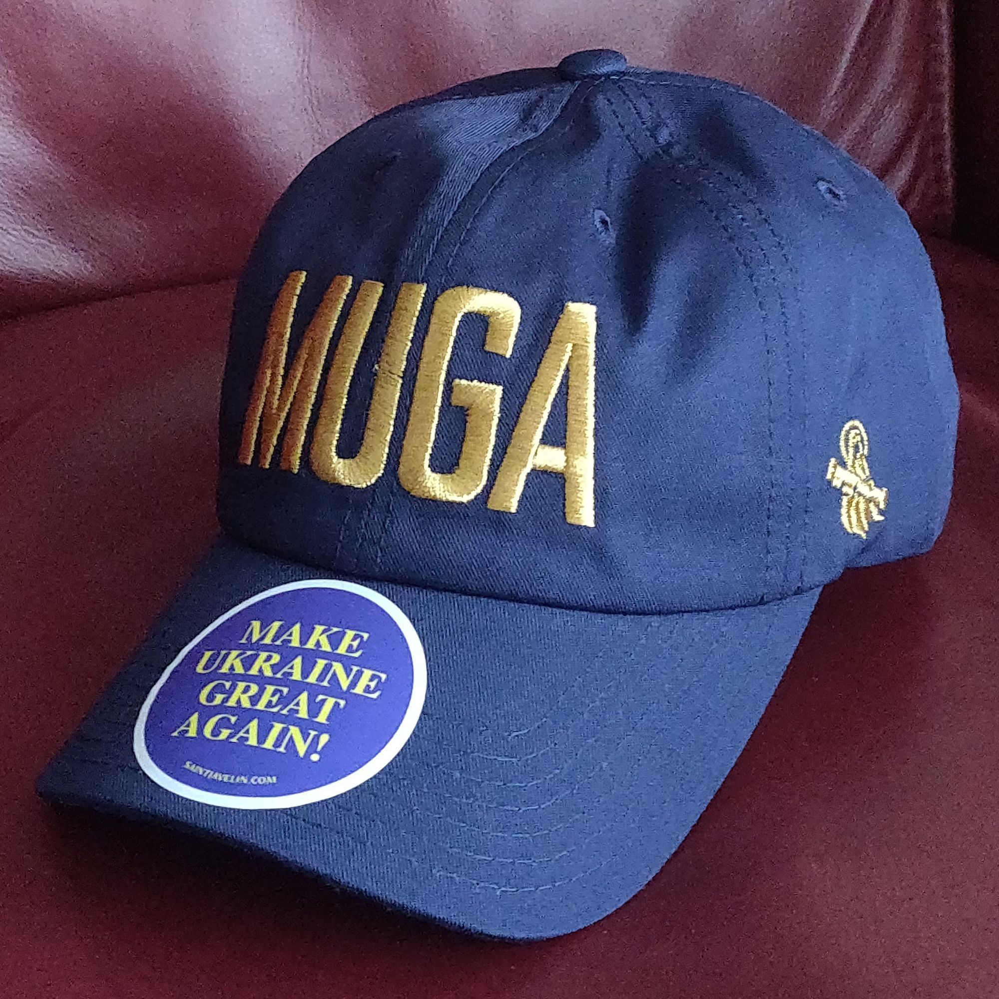 Blue hat, with gold-colored embroidered text "MUGA" on front and Saint Javelin logo on the side. On the bill there is a round sticker with the text "Make Ukraine Great Again".