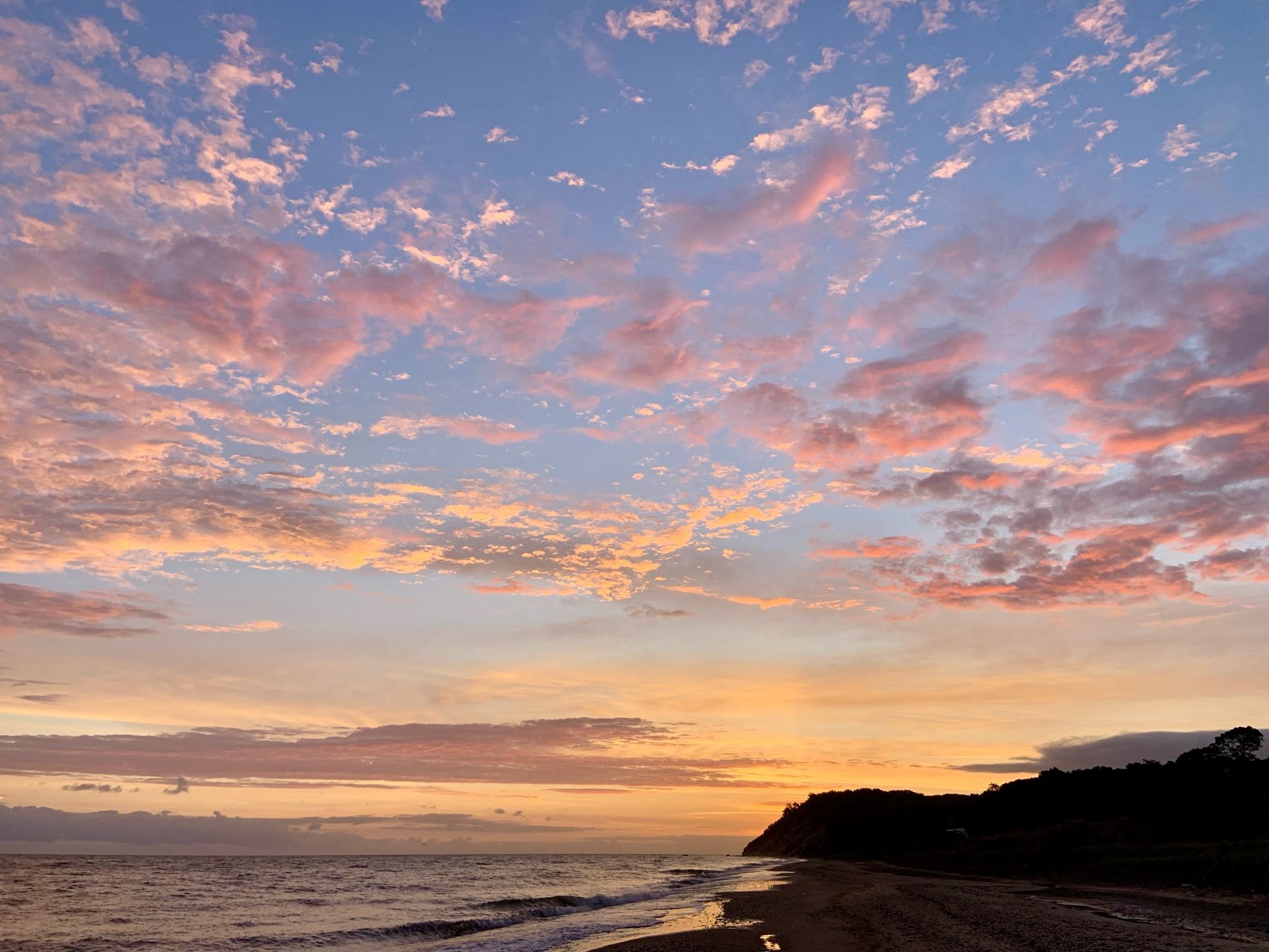 Photograph of a colorful sunrise above a beach. Bluffs mark the inland horizon.