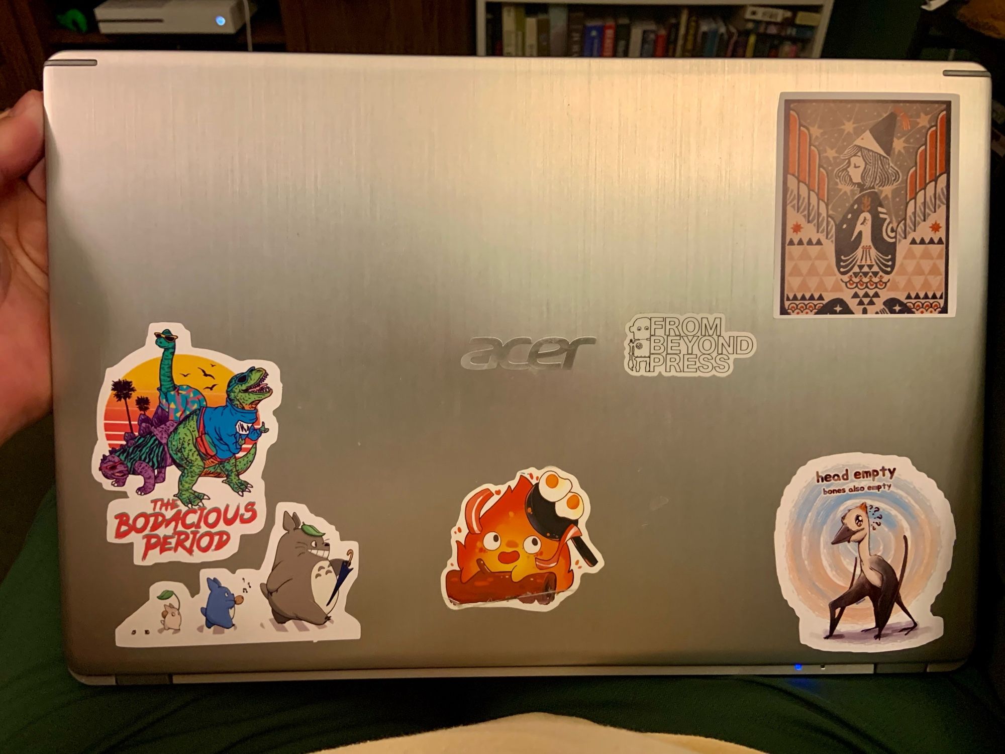 Photograph of my laptop, showing off various stickers I’ve applied to it. One is the logo of From Beyond Press. Others feature dinosaurs, Studio Ghibli characters, and a character from Witch Hat Atelier.