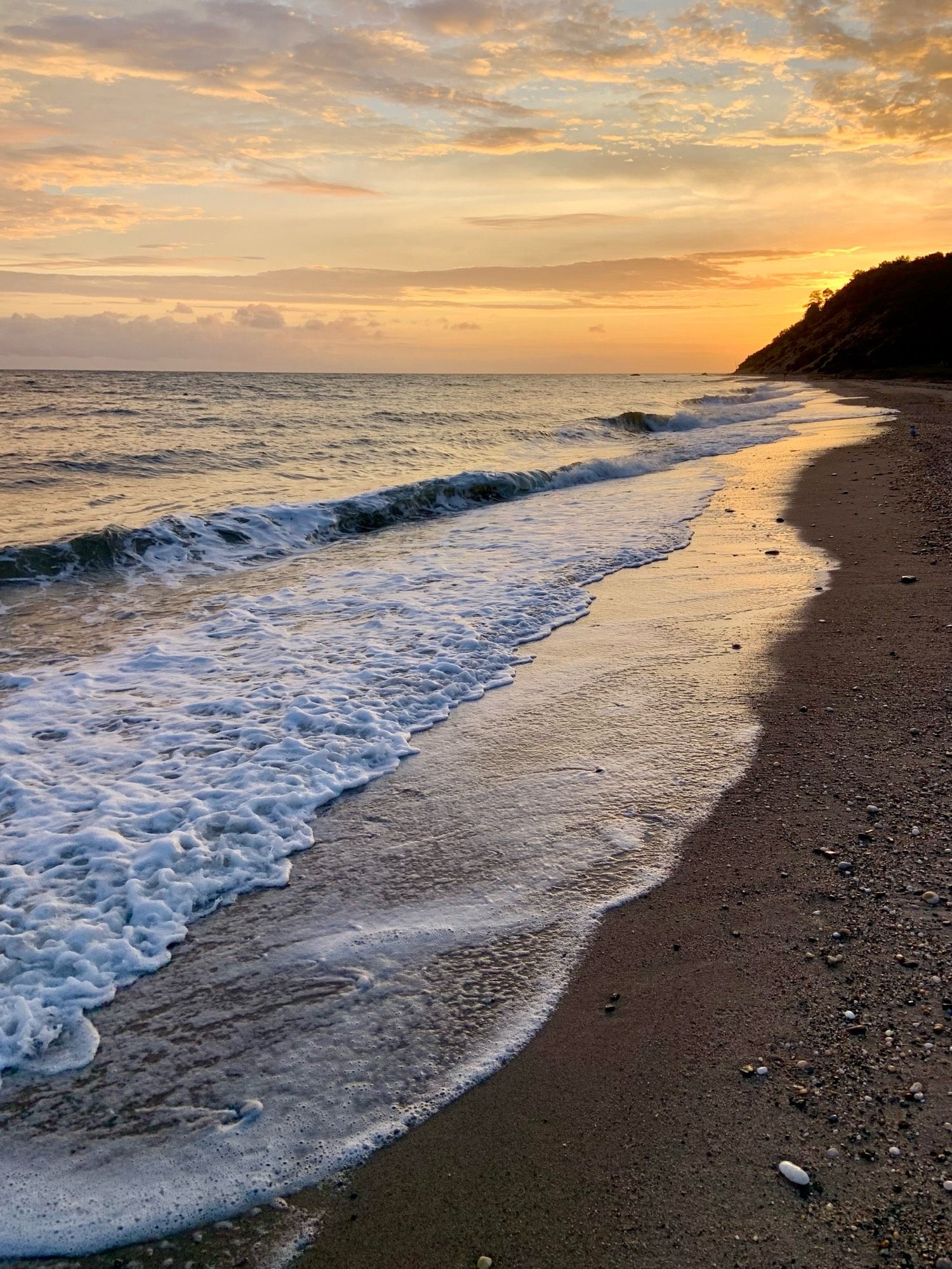 Photograph of sunrise on the beach. A line of low breakers marks the liminal edge between sea and sand.