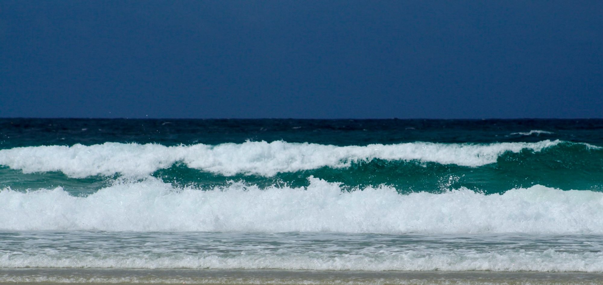 A photo of contrasts: a Prussian blue sky  against an almost black horizon, a strip of navy blue ocean, white foam crashing from turquoise blue waves, and a narrow band  of grey sea-battered sand.