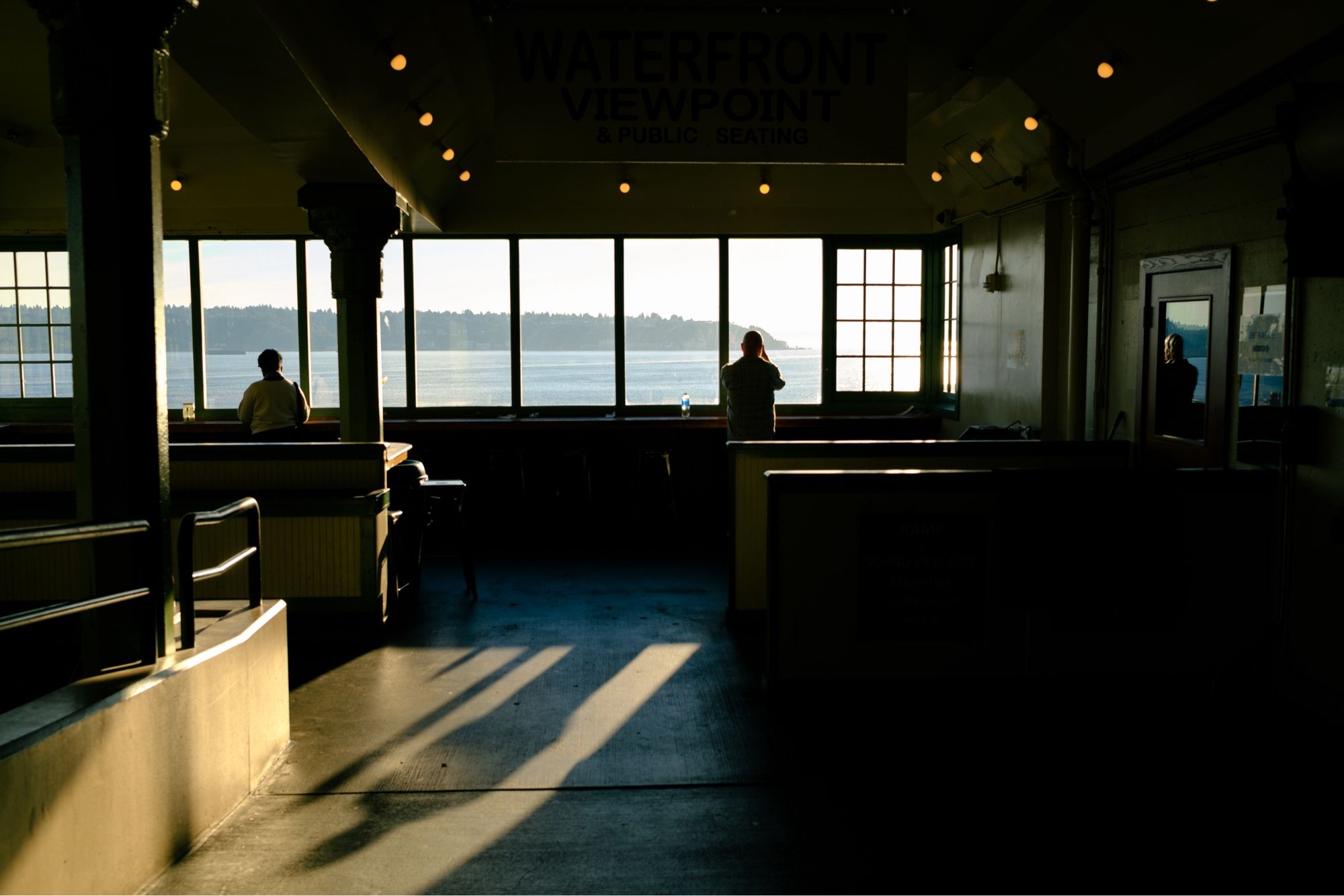 Two people back lit standing by a window with restaurant style booths near them, looking at the ocean beyond the glass