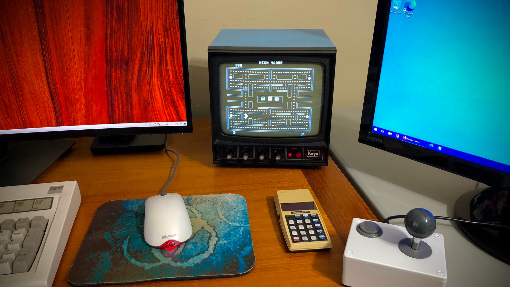 A photo of a desktop with two computer monitors. In between is a small B&W CRT security monitor showing Atari 800 Pac-Man. A joystick sits nearby.