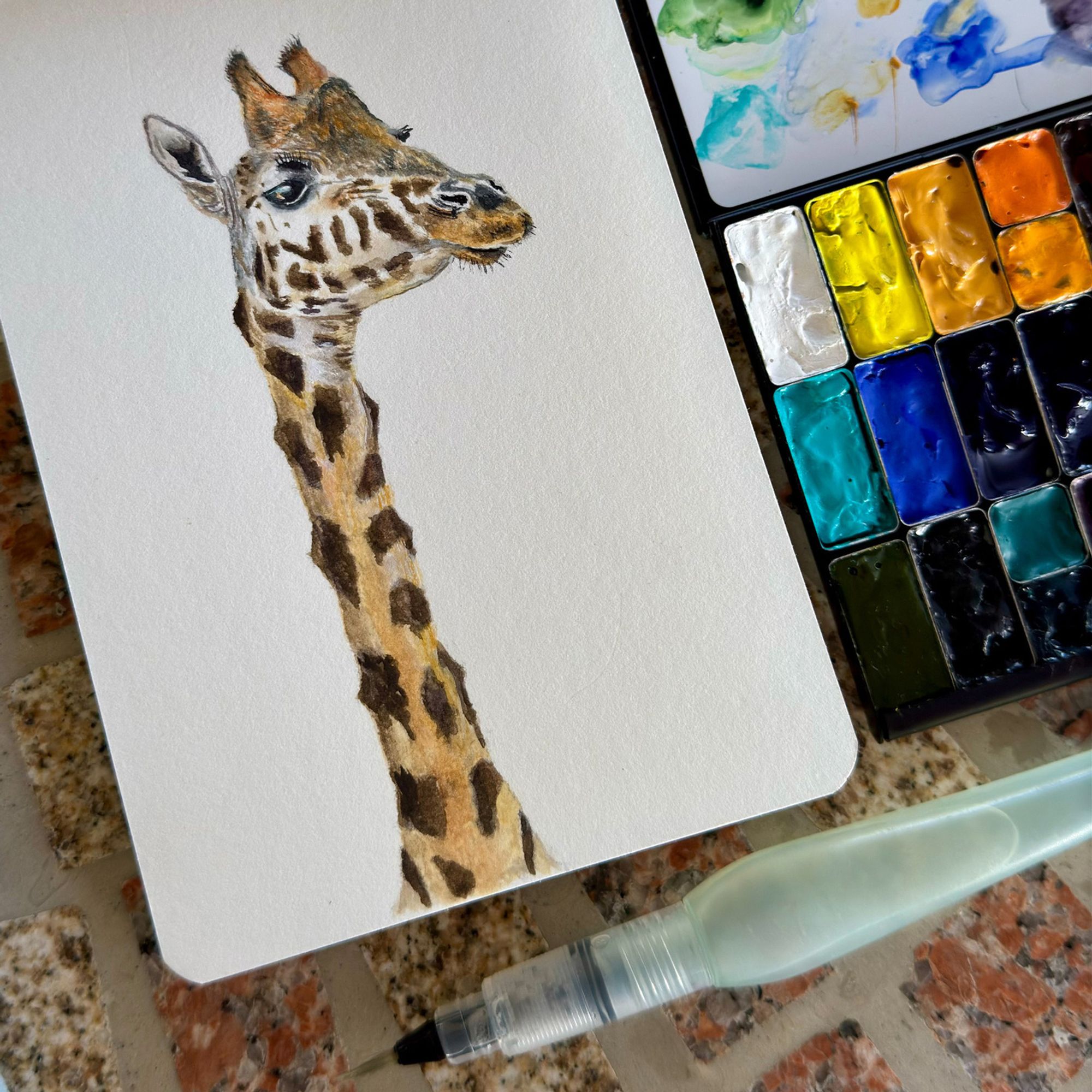 Watercolor painting of a giraffe's head and long neck, featuring a detailed pattern of spots. The painting is shown alongside a watercolor palette and a Pentel fine water brush. The artwork is done on a white sketchbook with smooth paper.