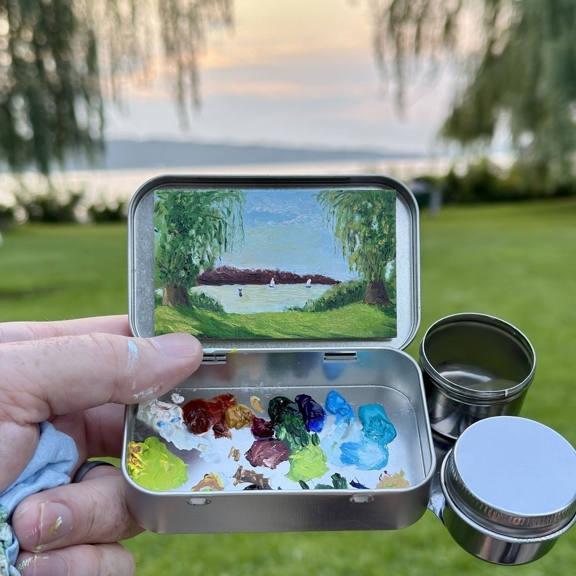 A small landscape painting in an open metal tin, held by an artist in front of a sunset scene at Stewart Park in Ithaca. The tin also contains vibrant oil paints in various colors. The painting captures trees, a distant island, and sailboats on a serene lake.&10;