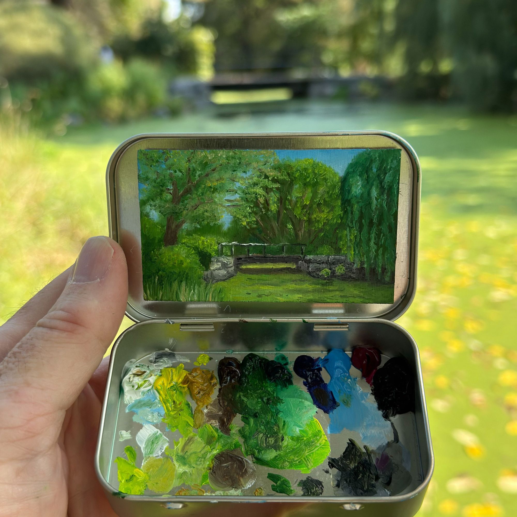 A tiny landscape painting inside a small metal tin, depicting a lush green scene with trees, shrubs, and a stone bridge. The tin also contains a palette with dabs of oil paint. The real landscape in the background matches the painting.&10;