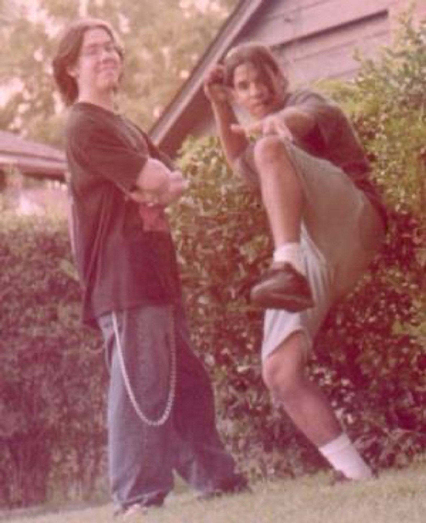 An over developed photo (hence the sepia color) of a normal white guy—author JD Buffington aged 17, c. 1997—and his high school best friend, normal Puerto Rican guy—anonymity protected—both posing for a camera. They both wear long black hair, JD’s dyed and wearing a blond streak on his left brow. JD wears overly baggy jeans and a chain that hangs below his knee from his belt to his wallet—as was the fashion at the time—and his friend is doing a faux karate pose. They stand in grass before bushes and a house.