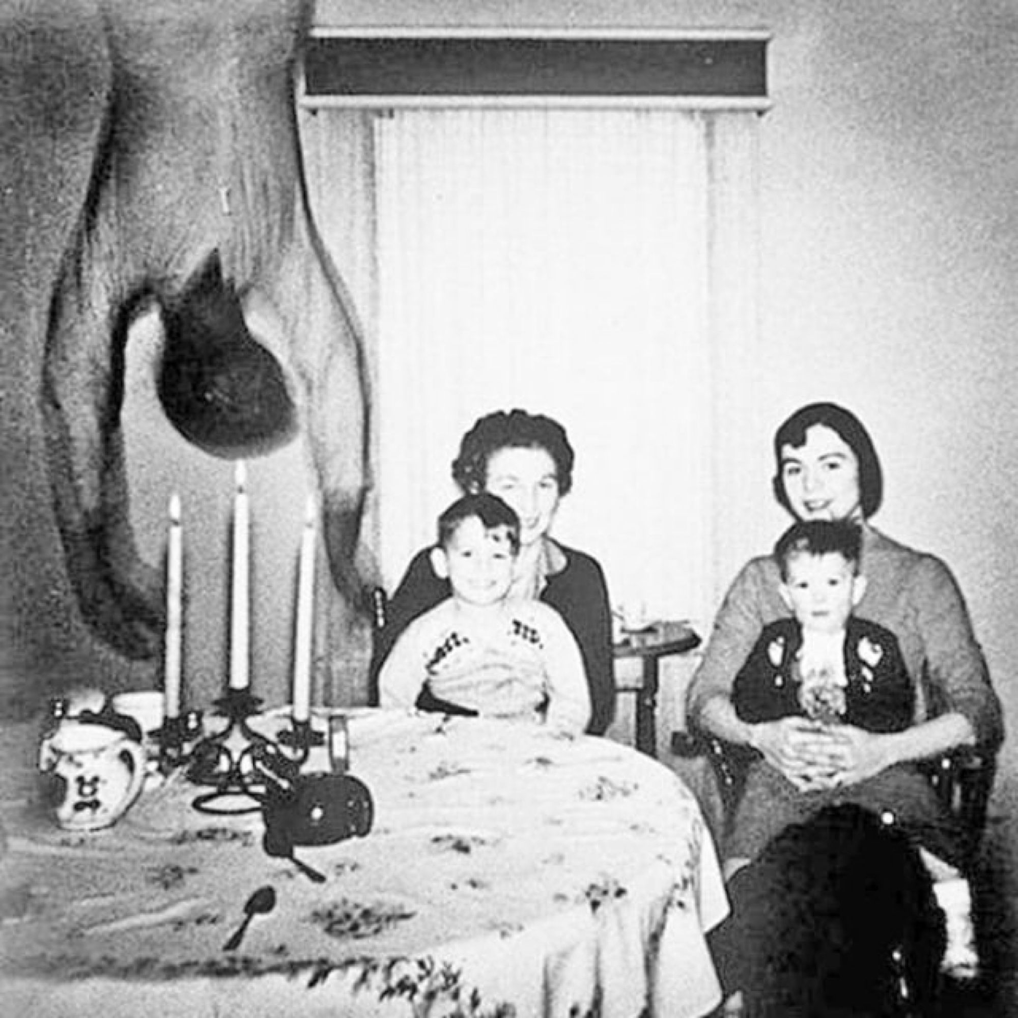 A black and white photo of two women sitting at a table with a child sitting in each of their laps. A disturbing figure hangs upside down from the top of the pic on the left side (to the women’s right) above the table and a candelabra. Its features are distorted and blurred as if in motion.