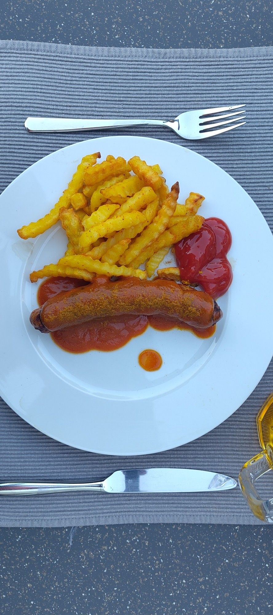 Eine Bratwurst mit Currysauce, Pommes Frittes und Ketchup auf einen runden weißen Porzellanteller. Daneben Messer und Gabel . Alles auf einem grauen Platzdeckchen.