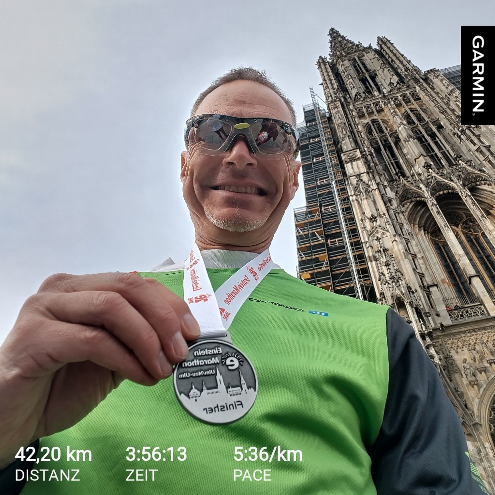 Ich mit Finisher Medaille Einsteinmarathon Ulm vor dem Ulmer Münster. Im Vordergrund Laufzeiten 42,2km 3:56:13 Zeit und entsprechende Pace