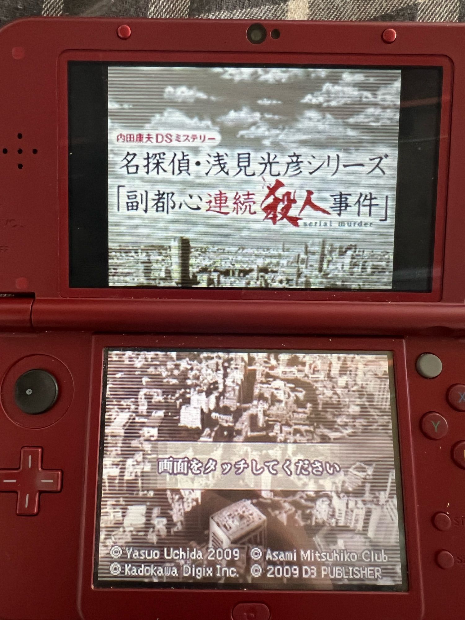 The title screen for the DS game 内田康夫DSミステリー名探偵・浅見光彦シリーズ「副都心連続殺人事件」 (Uchida Yasuo DS Mystery - Famous Detective Asami Mitsuhiko Series “The Fukutoshin Serial Murder Case”) Running on a red Nintendo New 3DS. The top screen has the game’s title and the bottom screen just says “Please touch the screen” in Japanese.