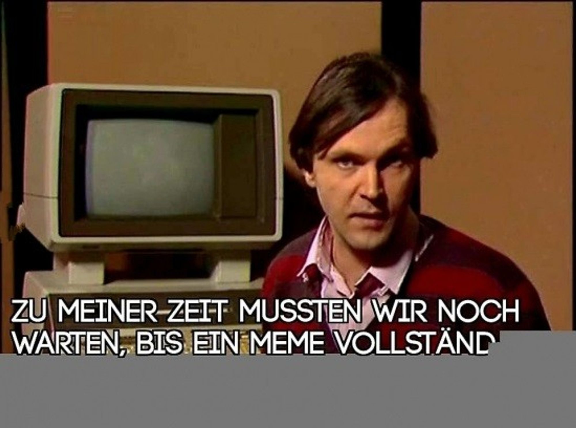 Das Bild zeigt eine Person, die vor einem altmodischen Computermonitor sitzt. Am unteren Rand des Bildes steht ein Text auf Deutsch: “Zu meiner Zeit mussten wir noch warten, bis ein Meme vollständ.”