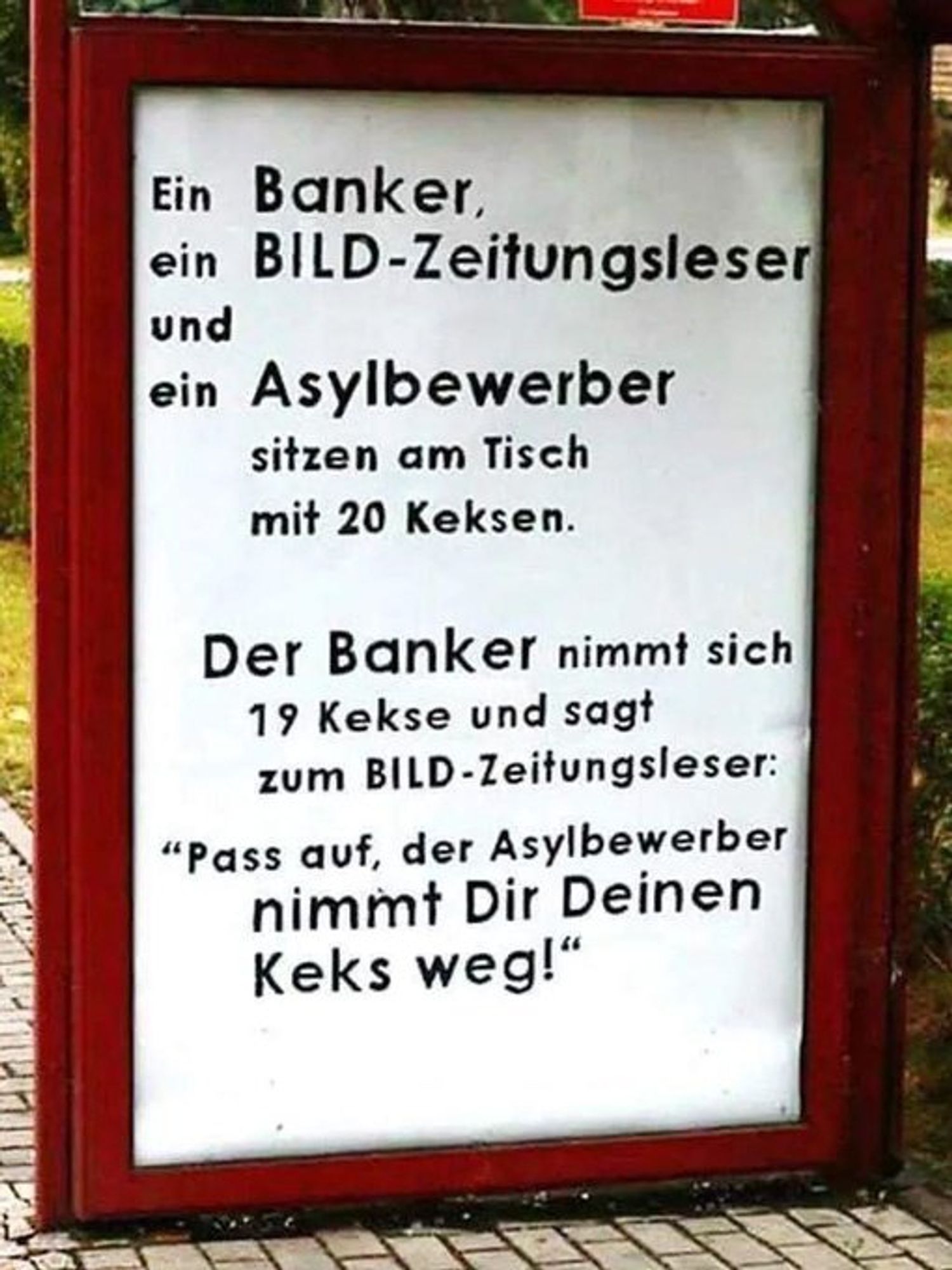 Ein gerahmtes Schild mit dem text:

Ein Banker,
ein BILD-Zeitungsleser
und
ein Asylbewerber
sitzen am Tisch
mit 20 Keksen.
Der Banker nimmt sich
19 Kekse und sagt
zum BILD-Zeitungsleser:
"Pass auf, der Asylbewerber
nimmt Dir Deinen
Keks weg!"