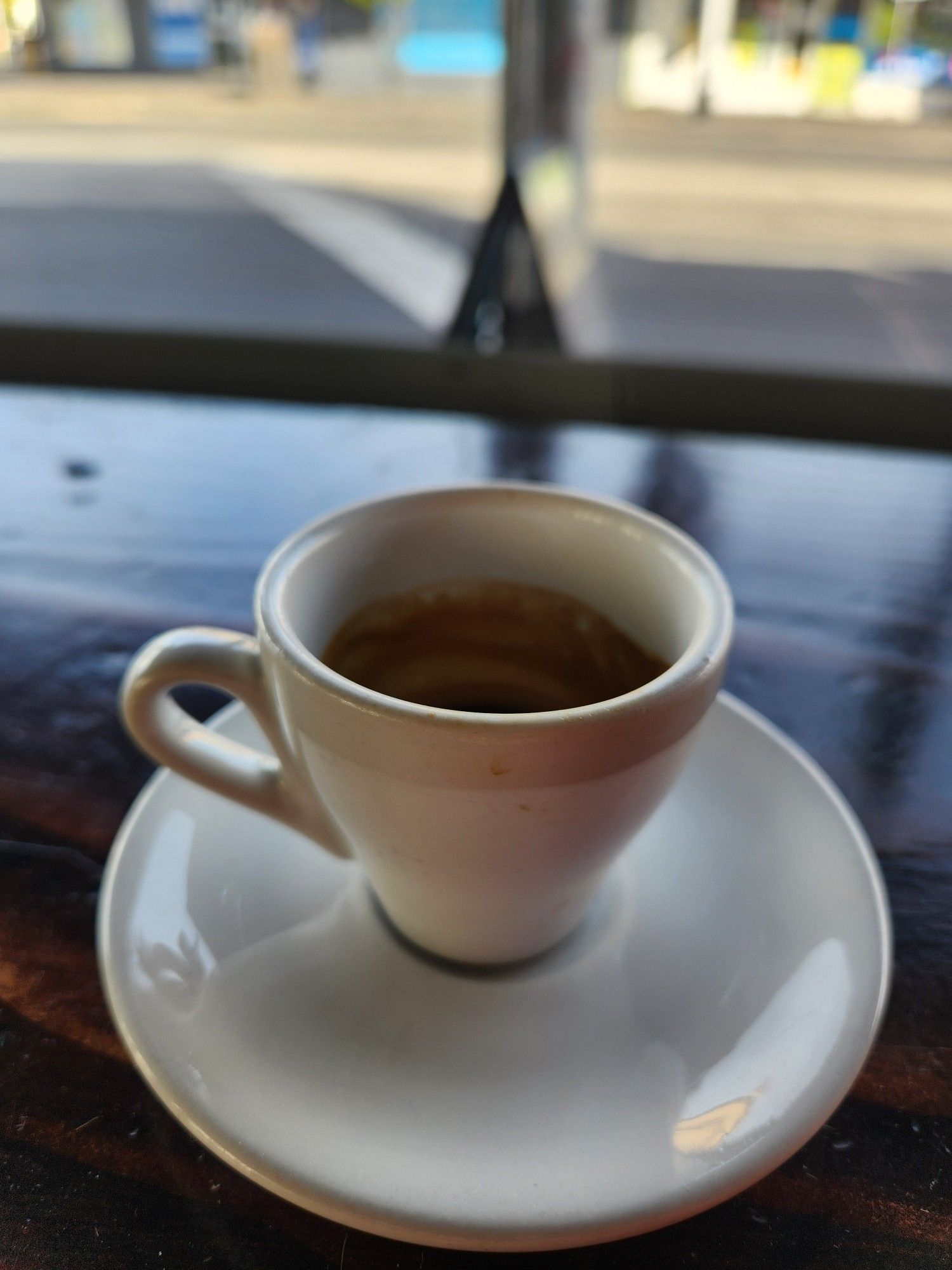 A white cup of espresso, in front of a window. The street outside is visible but out of focus.