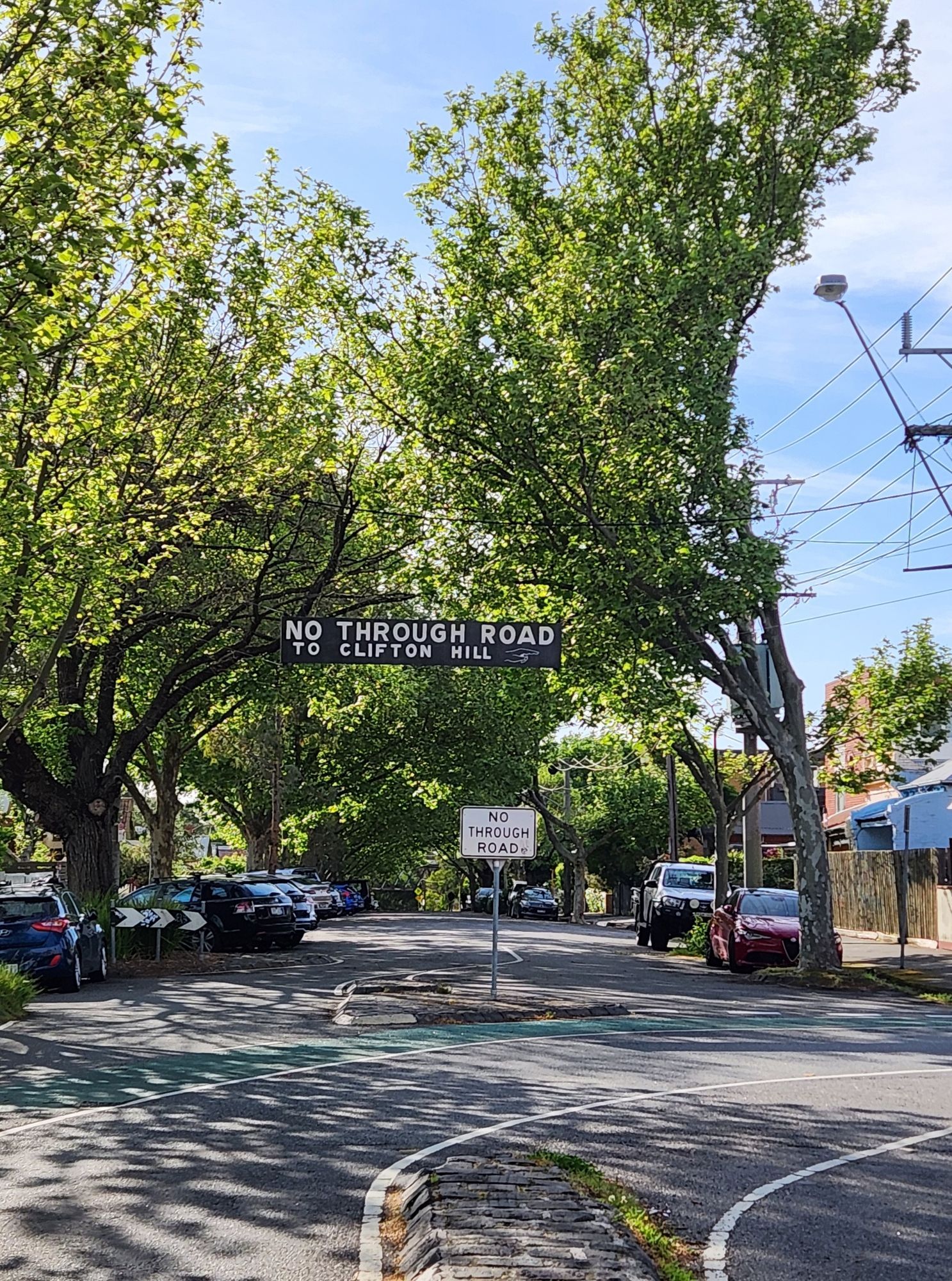 A quiet urban street planted with plane trees that form a canopy across the road. There is a white on black sign suspended above the road, reading "No through road to Clifton Hill".