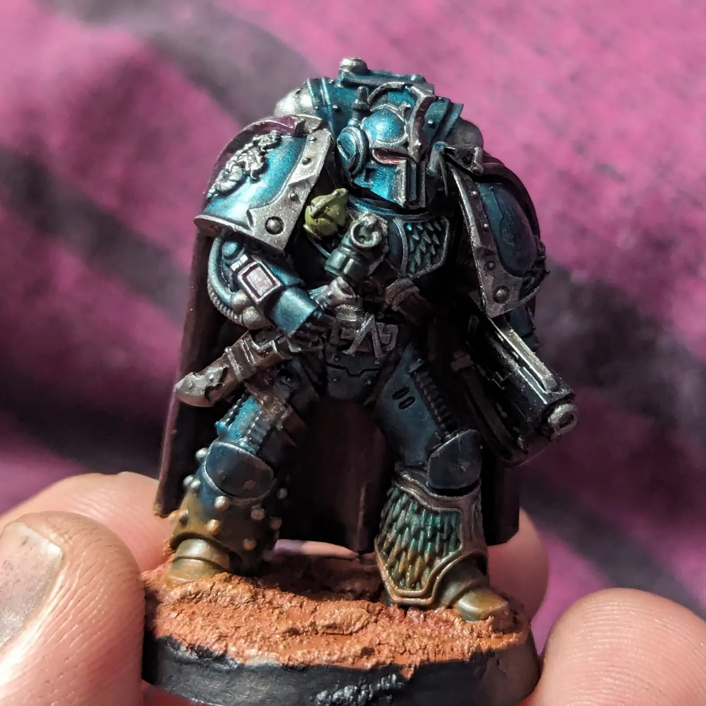 A space marine mini painted metallic blue with silver and metallic green accents gripping a combat knife and slinged rifle, equipped with several green grenades, wearing a purple cape, standing on a Marian base