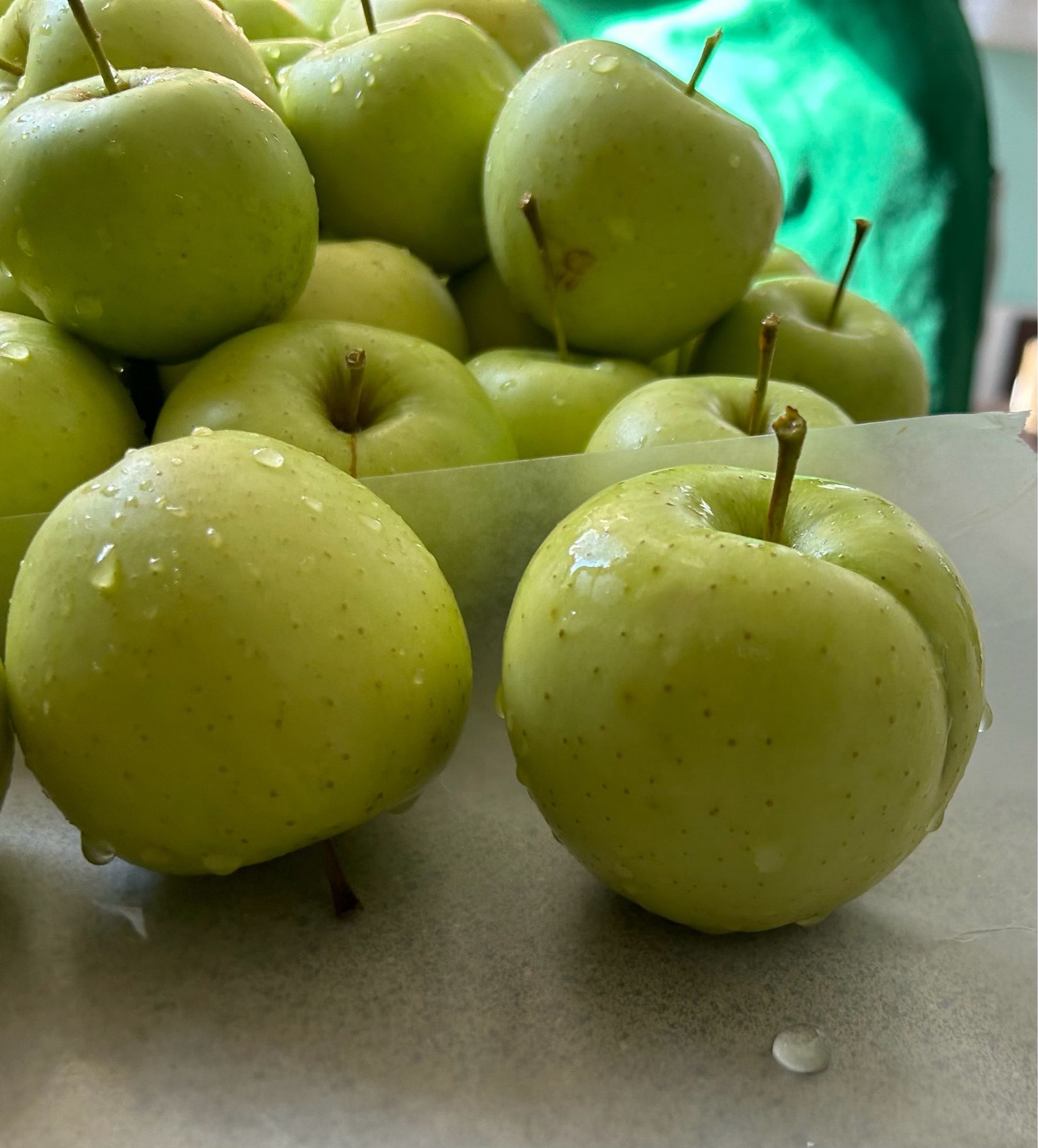 Several green apples cleaned and drying on a counter. The front apple has a crease or cleft that looks like a bottom.