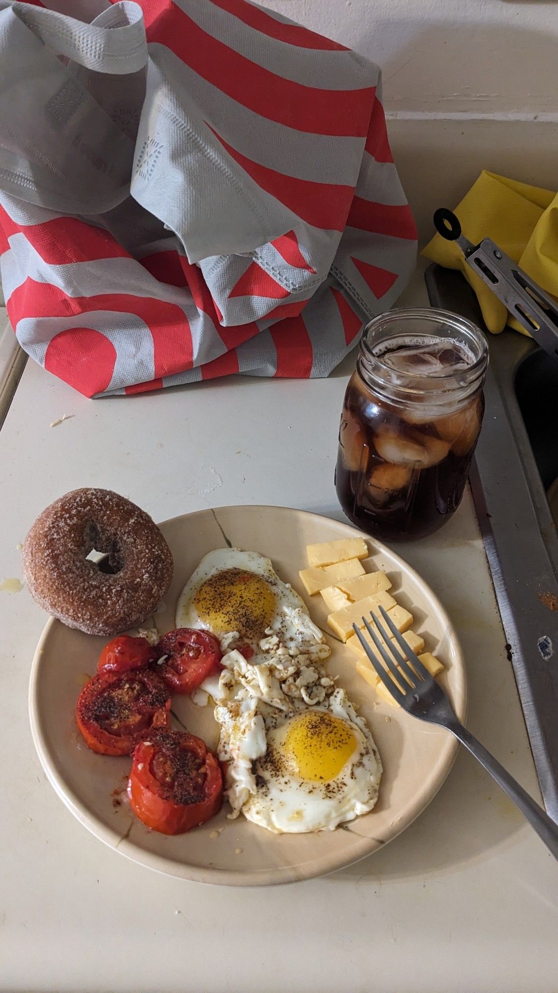 a cider donut, a glass of iced tea, two eggs sunny side up, some tomatoes, and slices  of gouda.