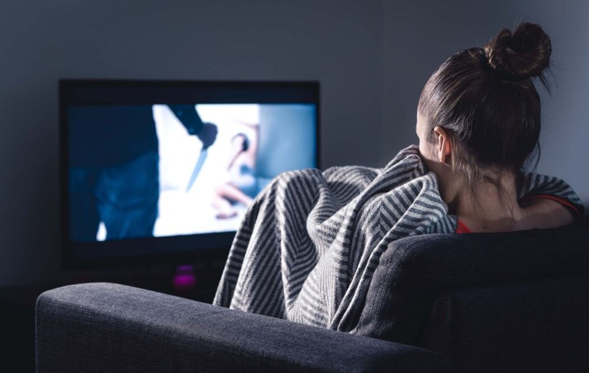 A person with dark hair tied in a bun sits on a grey couch, wrapped in a striped blanket. They are watching a suspenseful scene on TV, where a figure holds a knife, with a blurred view of a person in the background. The atmosphere appears tense, and the person watching is partially hiding the face with the blanket, suggesting they are frightened or anxious. The room is dimly lit, emphasising the glow from the TV screen,