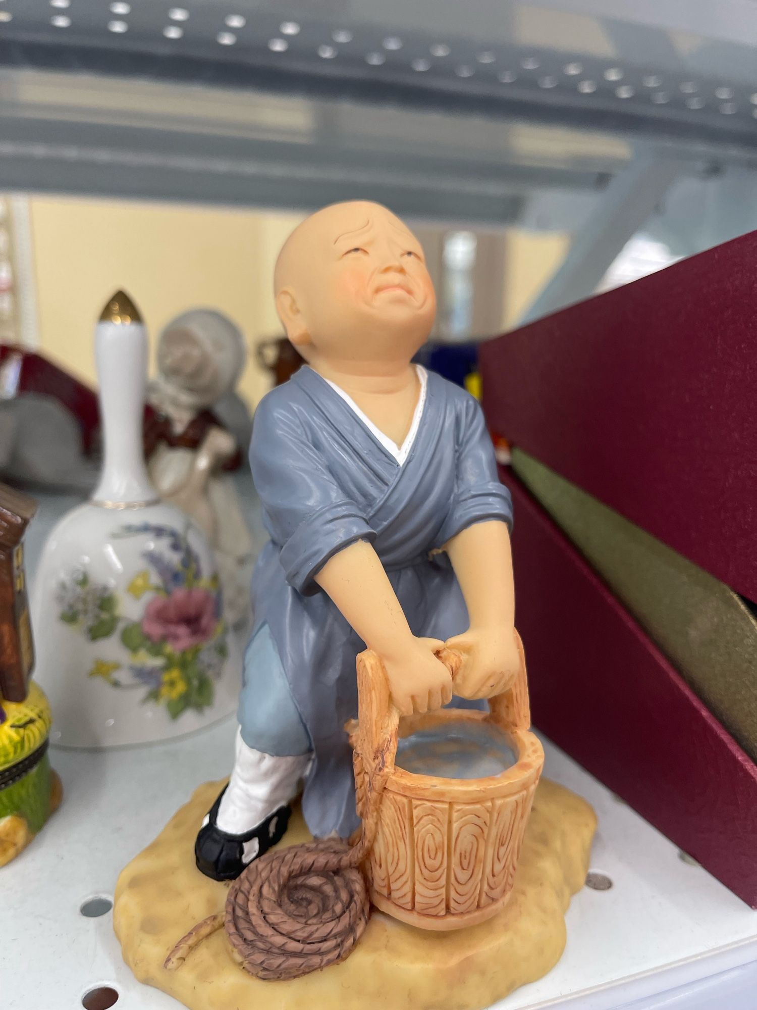 An approximately 6” figurine of of a chubby Asian child in ancient clothes, struggling with a bucket of water pulled up from a well. The subject looks like animated character Bobby Hill