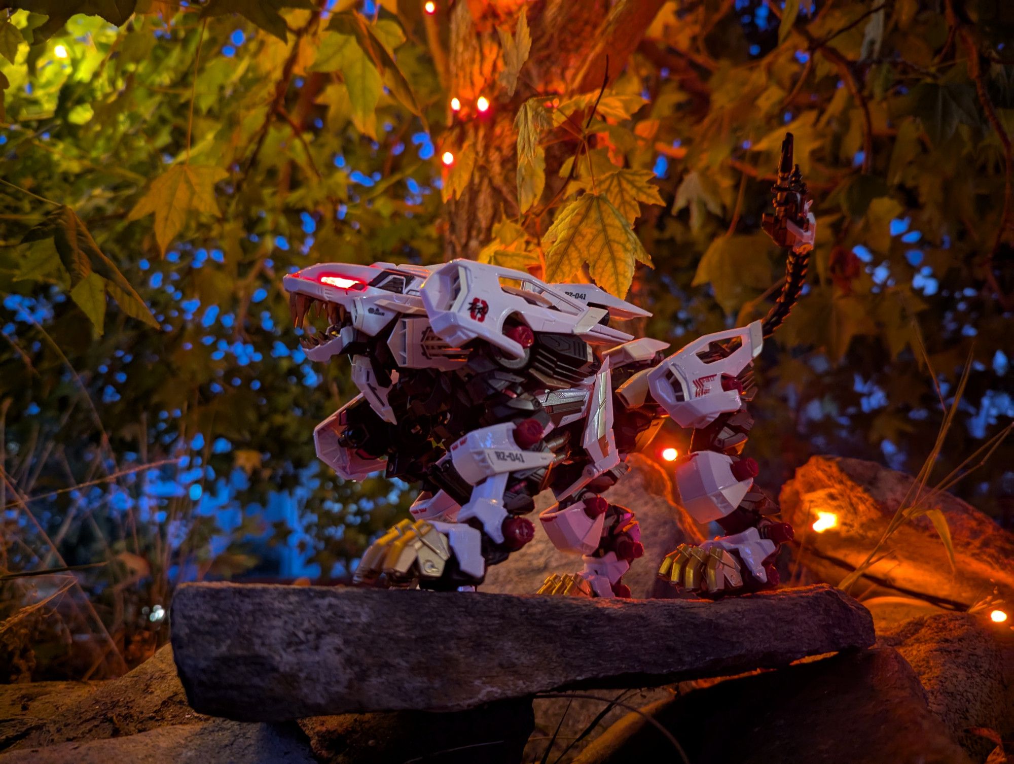 A photo of a Liger Zero from Zoids, posed on top of a stone head up in a roar and eyes glowing red. The photo is taken at dusk and lit with orange holiday lights, while the background is made of patches of dark blue sky showing through green and yellow leaves of a tree.