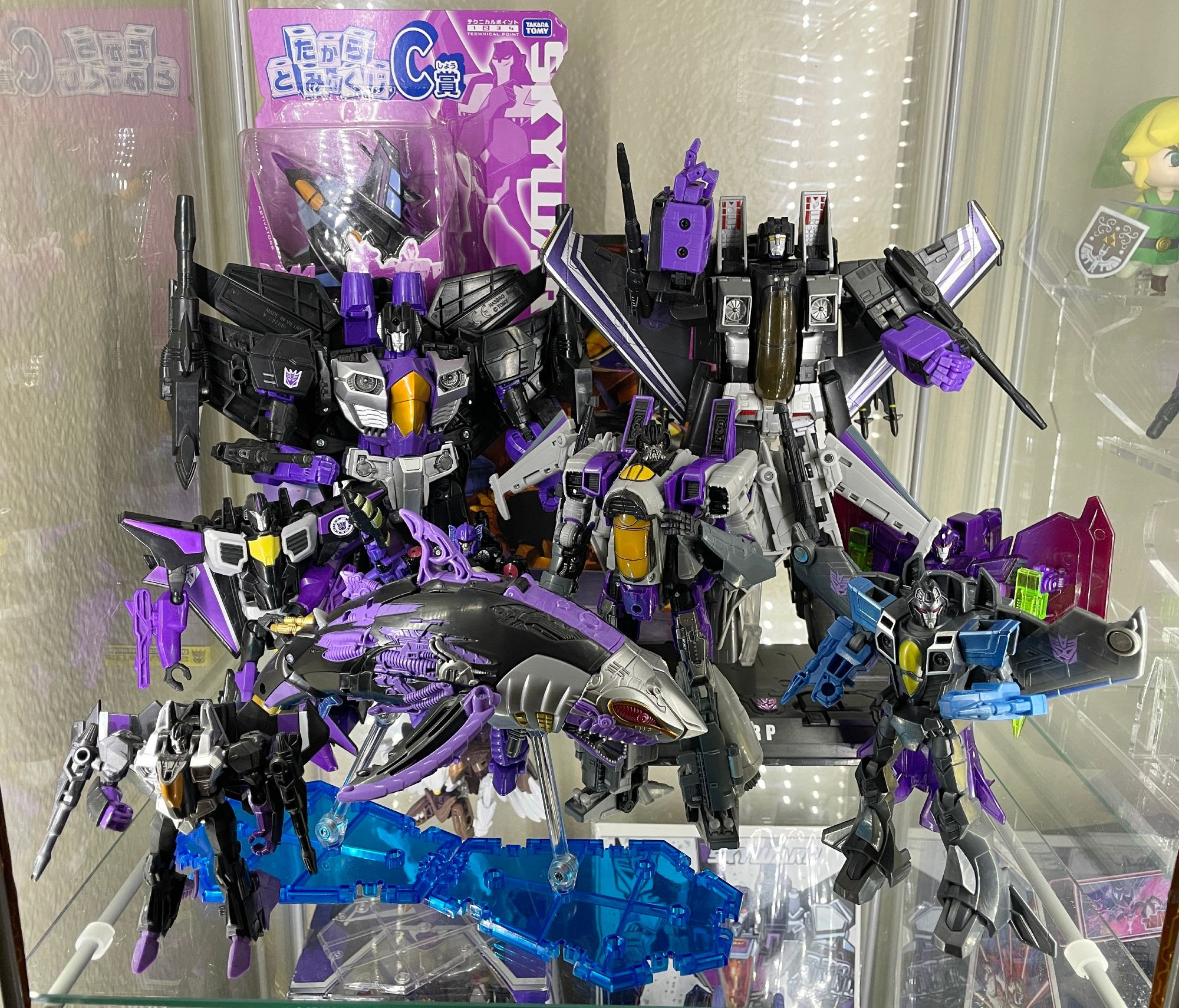 A display shelf full of Transformers Skywarp figures. The old Masterpiece figure, Combiner Wars, the Bumblebee movie, Robots in Disguise, Cyberverse, Earthspark, the "Hellwarp" shark version of him, and a couple more.